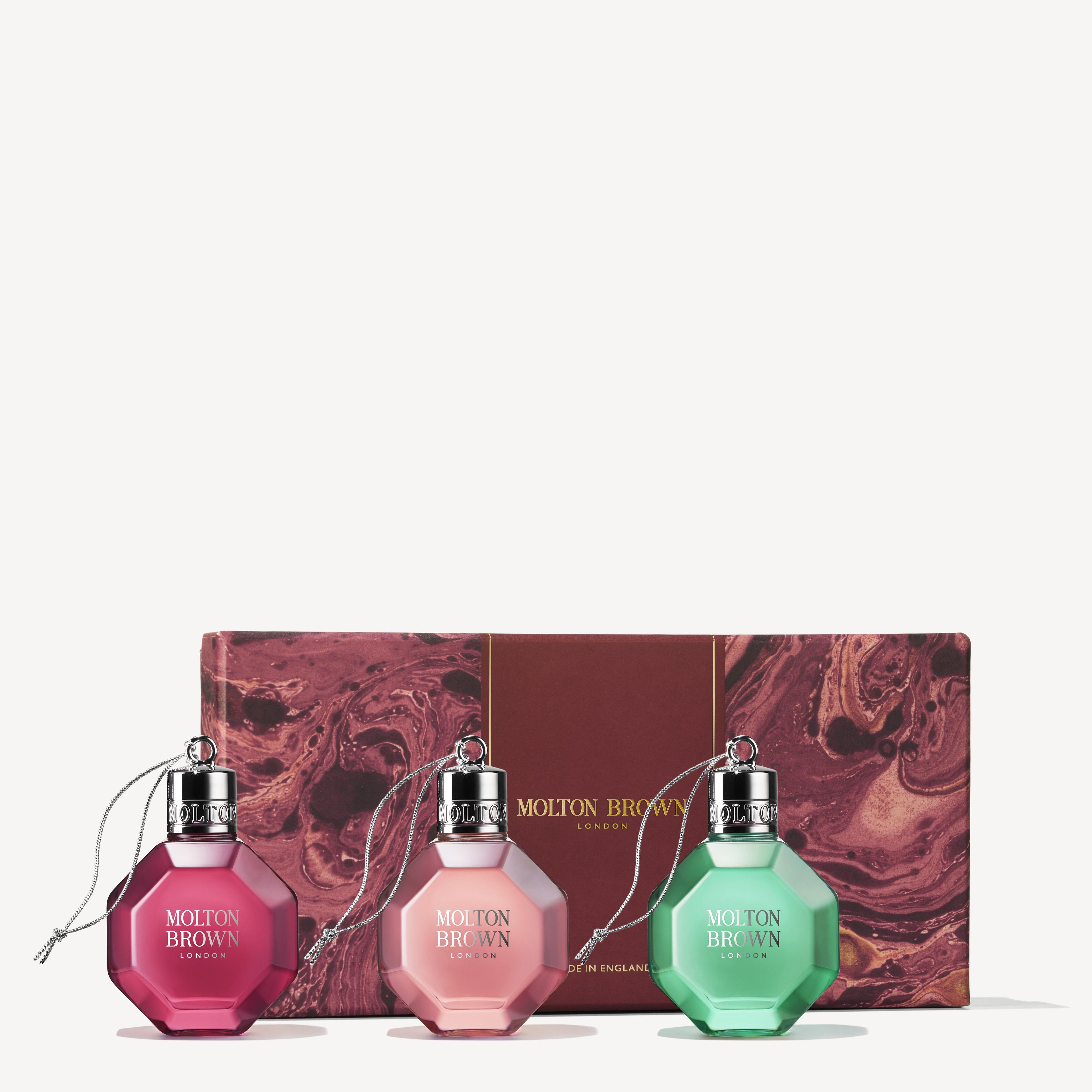 A Festive Bauble Gift Set from MoltonBrown Cyprus, featuring three vibrant baubles filled with aromatic, fruity perfumes, is elegantly displayed in front of a decorative marbled gift box. The baubles are in shades of red, pink, and green, each adorned with a silver cap and string for hanging.