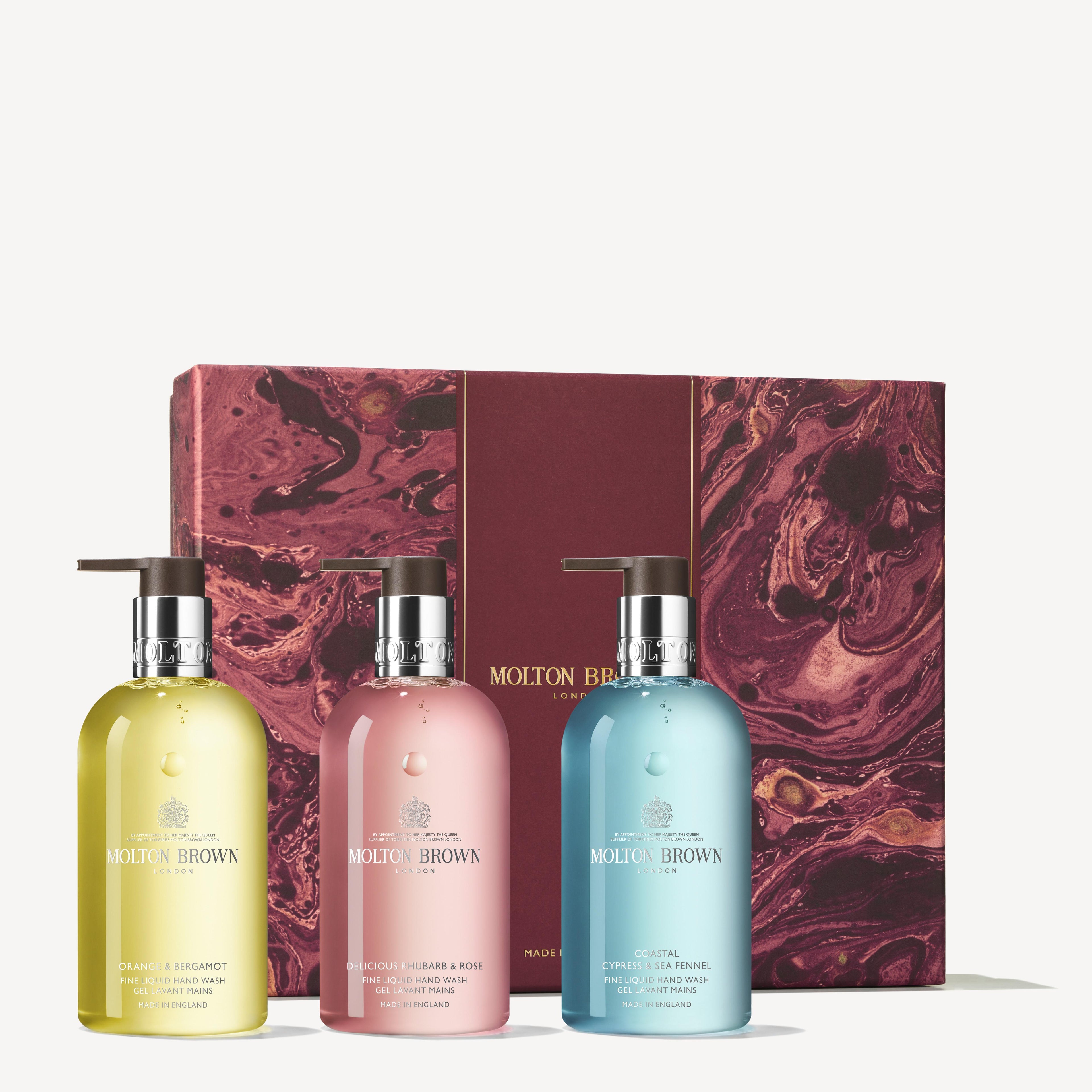 Three MoltonBrown Cyprus bottles in yellow, pink, and blue are arranged in front of a maroon gift box adorned with gold swirls. Featuring the Floral & Aromatic Hand Care Collection, these bottles have pump dispensers and prominently display the brand name. They perfectly exemplify citrus floral-fruity aromatic scents.