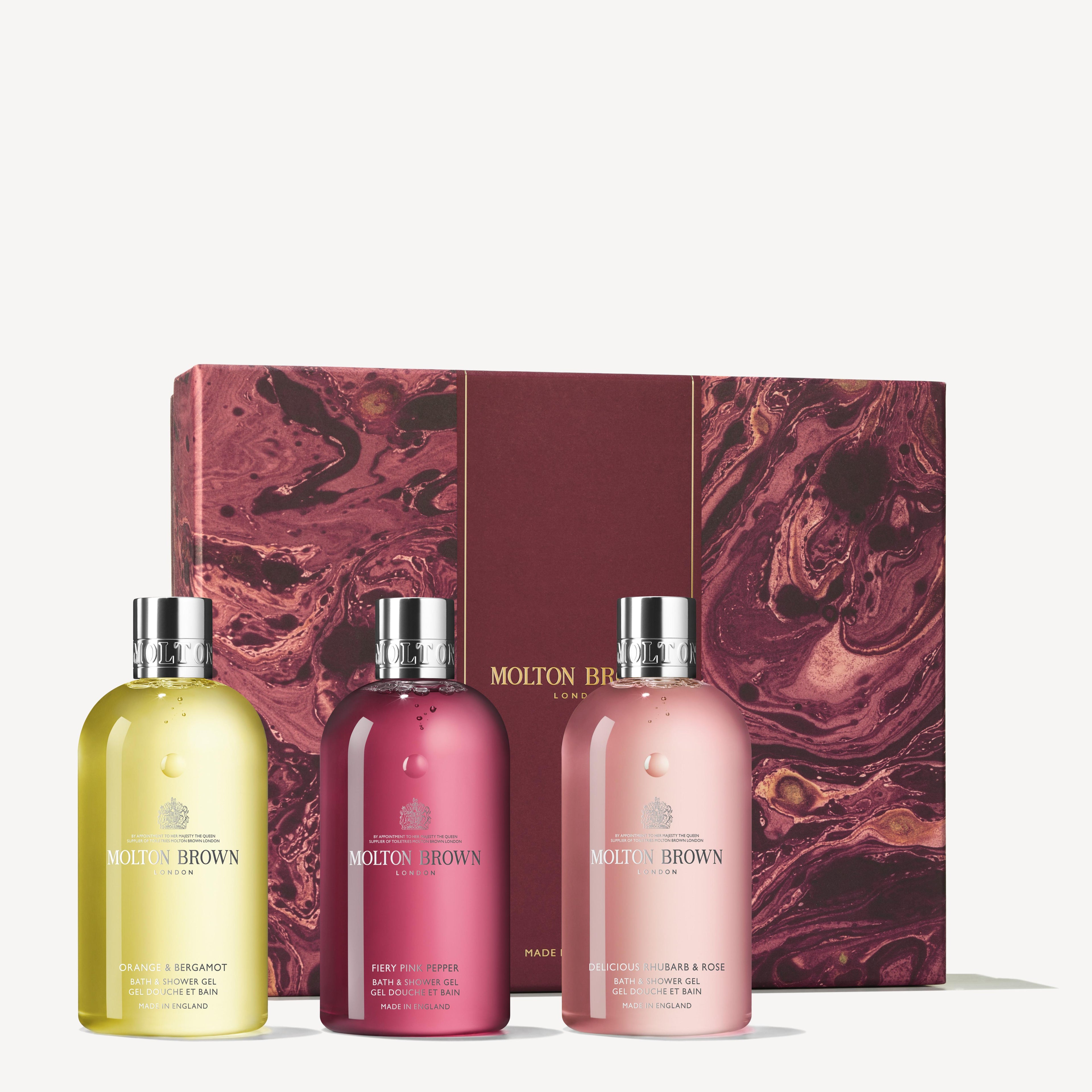 Three bottles from the MoltonBrown Cyprus Floral & Spicy Body Care Gift Set are elegantly positioned in front of a marbled red and pink gift box. The bottles, displaying a spectrum of colors—yellow, pink, and red—and topped with silver caps, stand out against a light cream background that emphasizes the exquisite fragrance concentrations within these luxurious body washes.