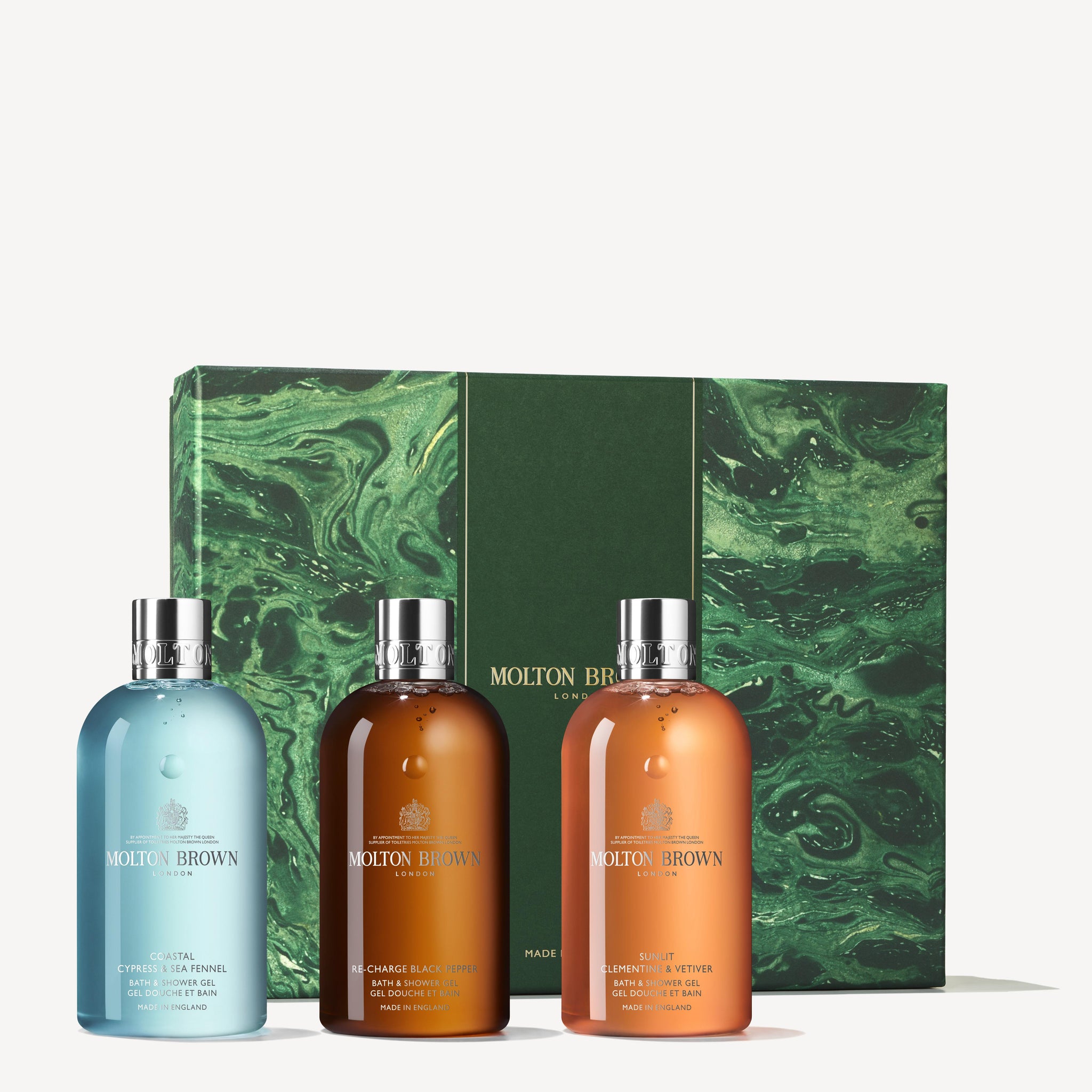 A set from MoltonBrown Cyprus, called the Woody & Aromatic Body Care Gift Set, includes three bottles adorned in striking blue, brown, and orange hues. Each bottle exudes a captivating aromatic fragrance. The backdrop displays an elegant dark green gift box with a marble pattern, showcasing the MoltonBrown logo and text—ideal for a winter cleansing ritual.