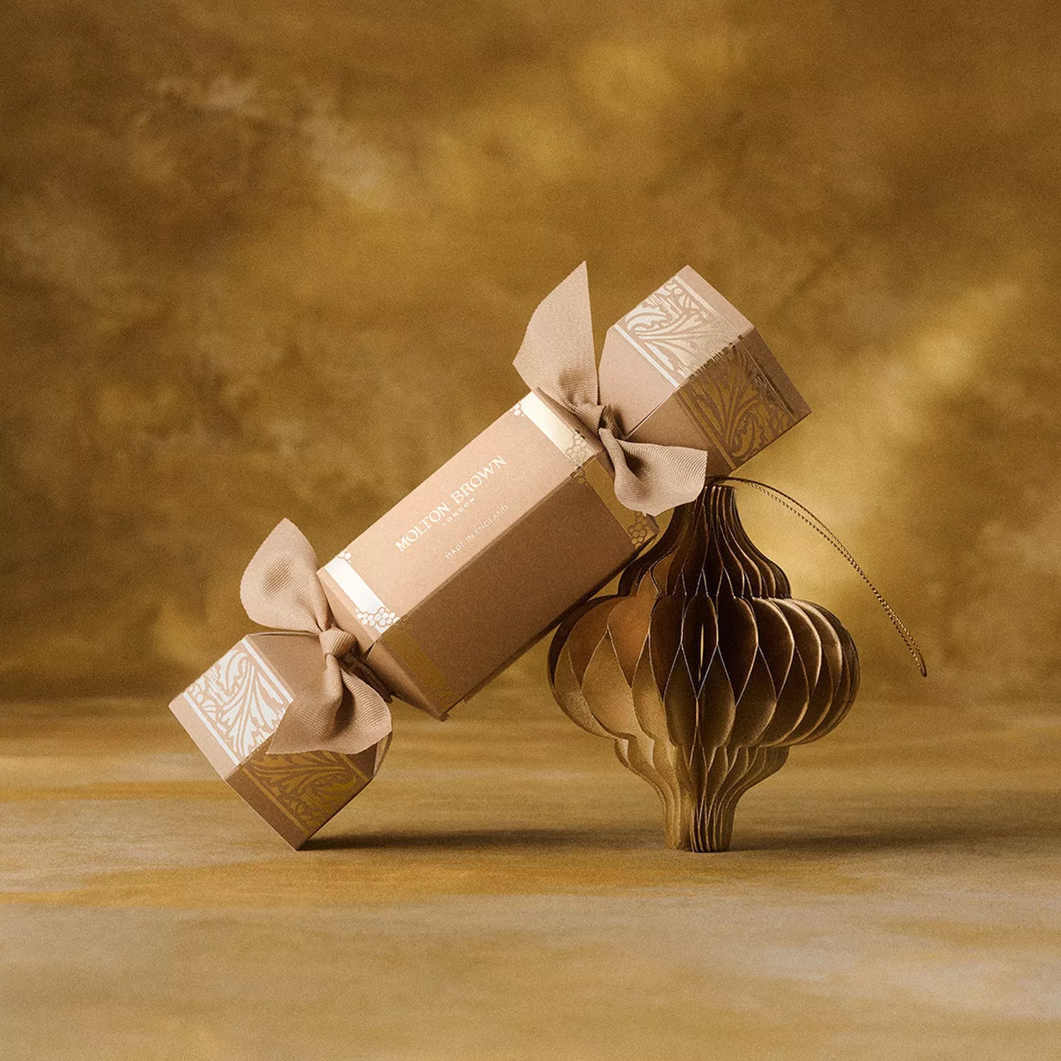 The MoltonBrown Cyprus Orange & Bergamot Christmas Cracker, tied with ribbon bows, rests atop a gold ornament against a warm backdrop—an ideal sparkling table gift for the festive season.