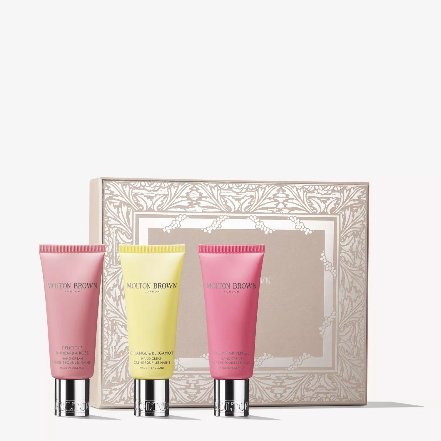 Three vibrant MoltonBrown Cyprus Floral & Spicy Hand Cream tubes stand before a decorative beige gift box with ornate white borders, each enriched with classic fragrances and nourishing shea butter.