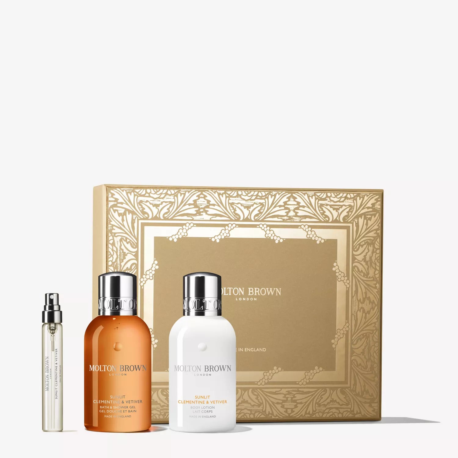 The Sunlit Clementine & Vetiver Travel Gift Set by MoltonBrown Cyprus features a gold box with an ornate floral border, holding three products: citrus perfume travel spray, orange body wash, and fig leaf lotion—all labeled Clementine & Vetiver.