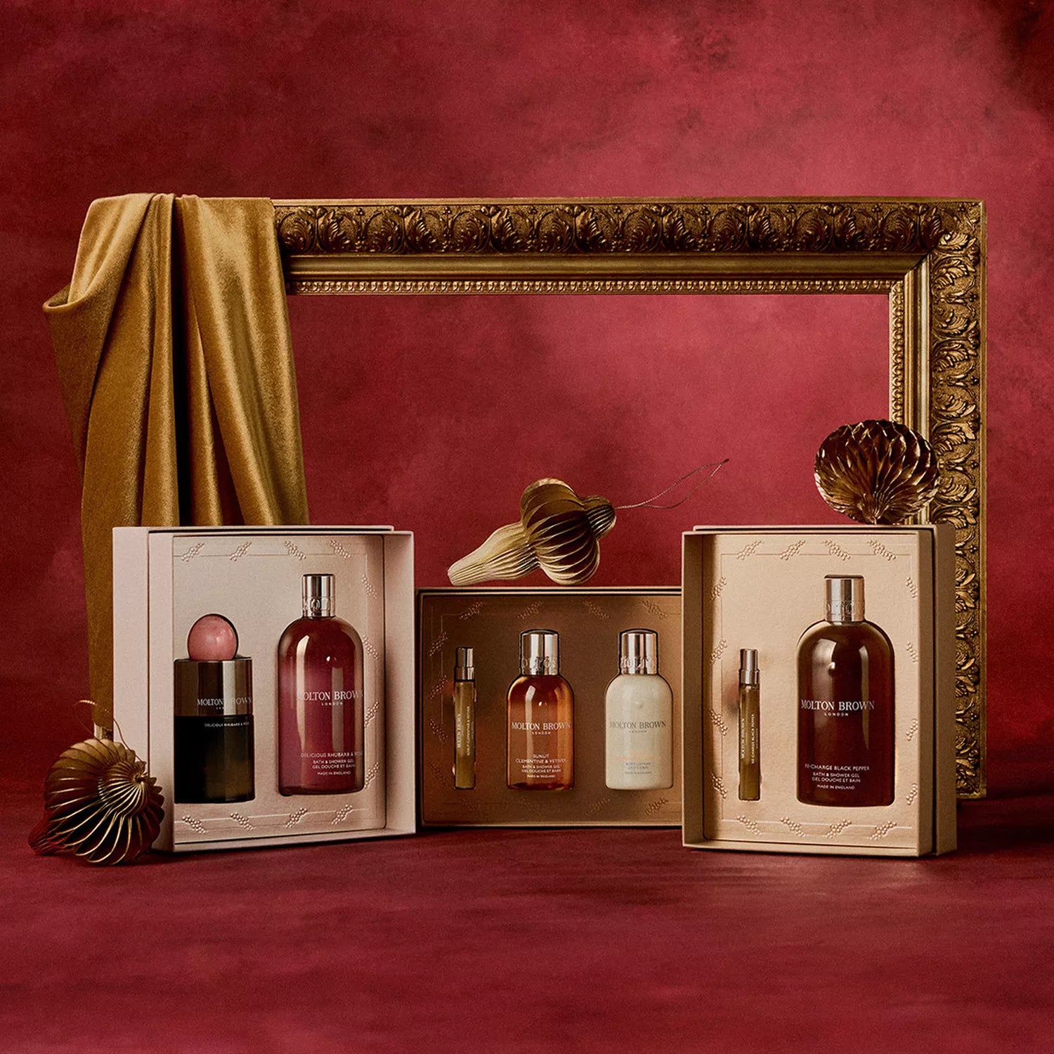 Three luxurious MoltonBrown Cyprus Sunlit Clementine & Vetiver Travel Gift Sets with body wash and bath products sit on red velvet, framed by a gold picture frame and fabric, with paper fans adding decorative flair.