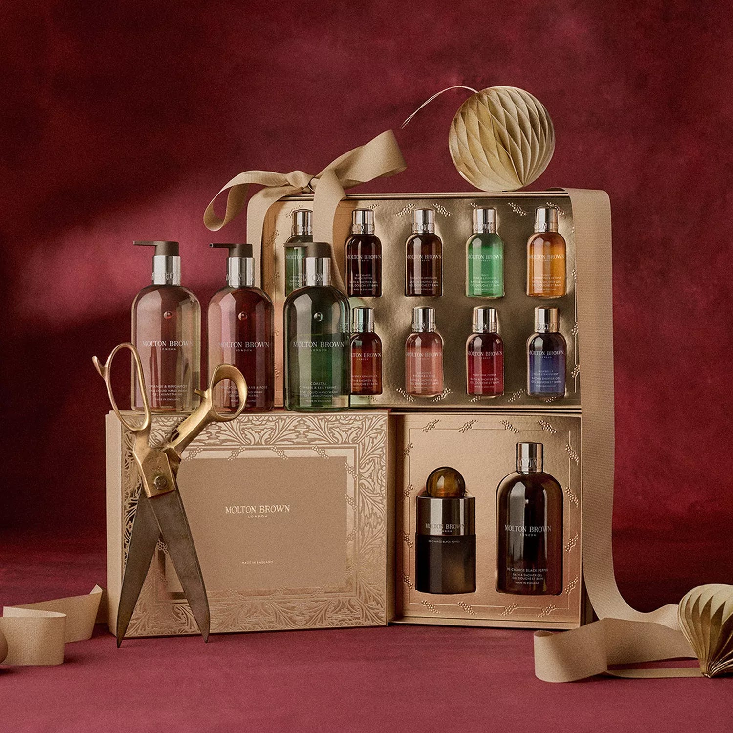 The MoltonBrown Cyprus Stocking Filler Gift Set features body wash, hand wash, and mini bath & shower gels with large gold scissors, ribbon, and gold accents—all elegantly arranged on red velvet.