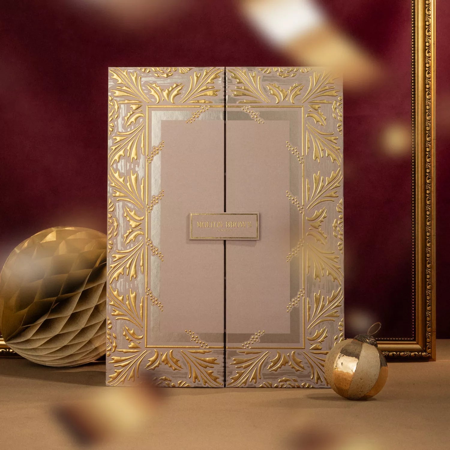 A luxurious light gold gift box with gold leaf designs stands upright against a burgundy background. The Advent Calendar by MoltonBrown Cyprus promises scented treasures inside, with a gold ornament and honeycomb decoration nearby.