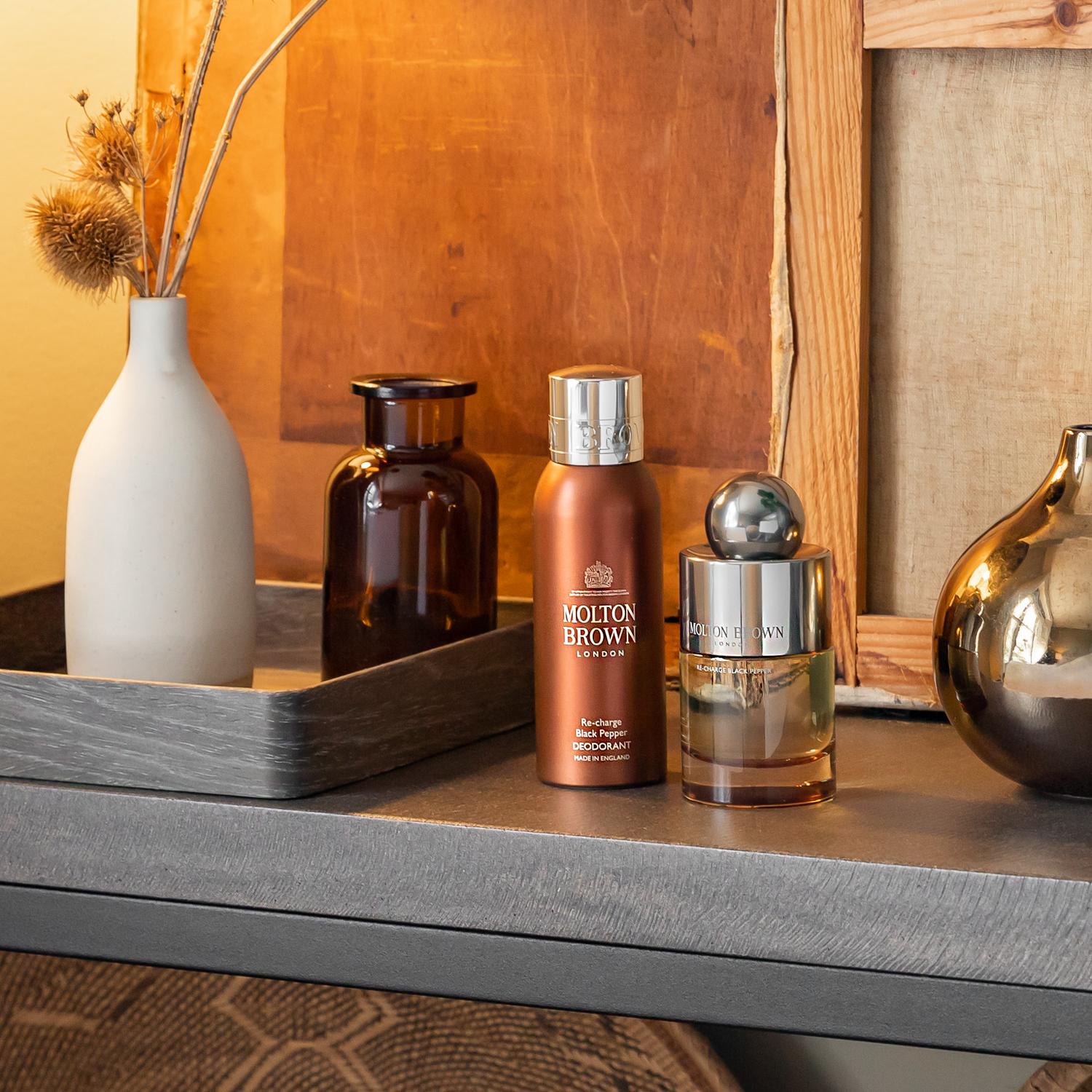 A decorative arrangement showcases a Re-charge Black Pepper Deodorant 150ml from MoltonBrown Cyprus alongside a Molton Brown perfume bottle on a gray shelf, emanating an iconic scent and long-lasting freshness. A white vase with dried flowers and a brown vase accompany them against wooden frames in the background.