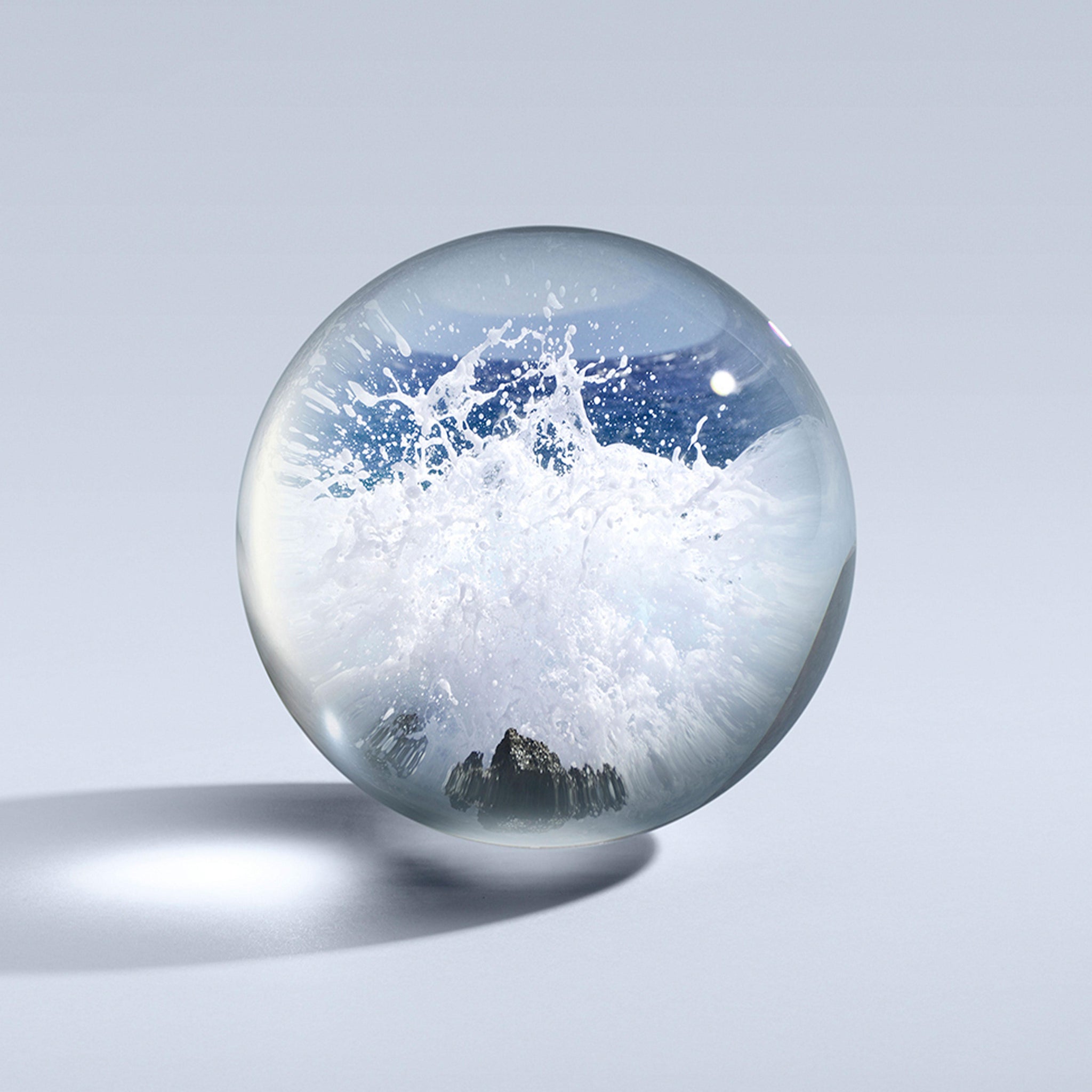 A crystal-clear orb rests on a smooth surface, capturing a lively tableau of ocean waves crashing against rocks, with notes reminiscent of the scent of MoltonBrown Cyprus' Coastal Cypress & Sea Fennel Deodorant. It feels like holding a miniature ocean sphere surrounded by a gentle, neutral gray setting.