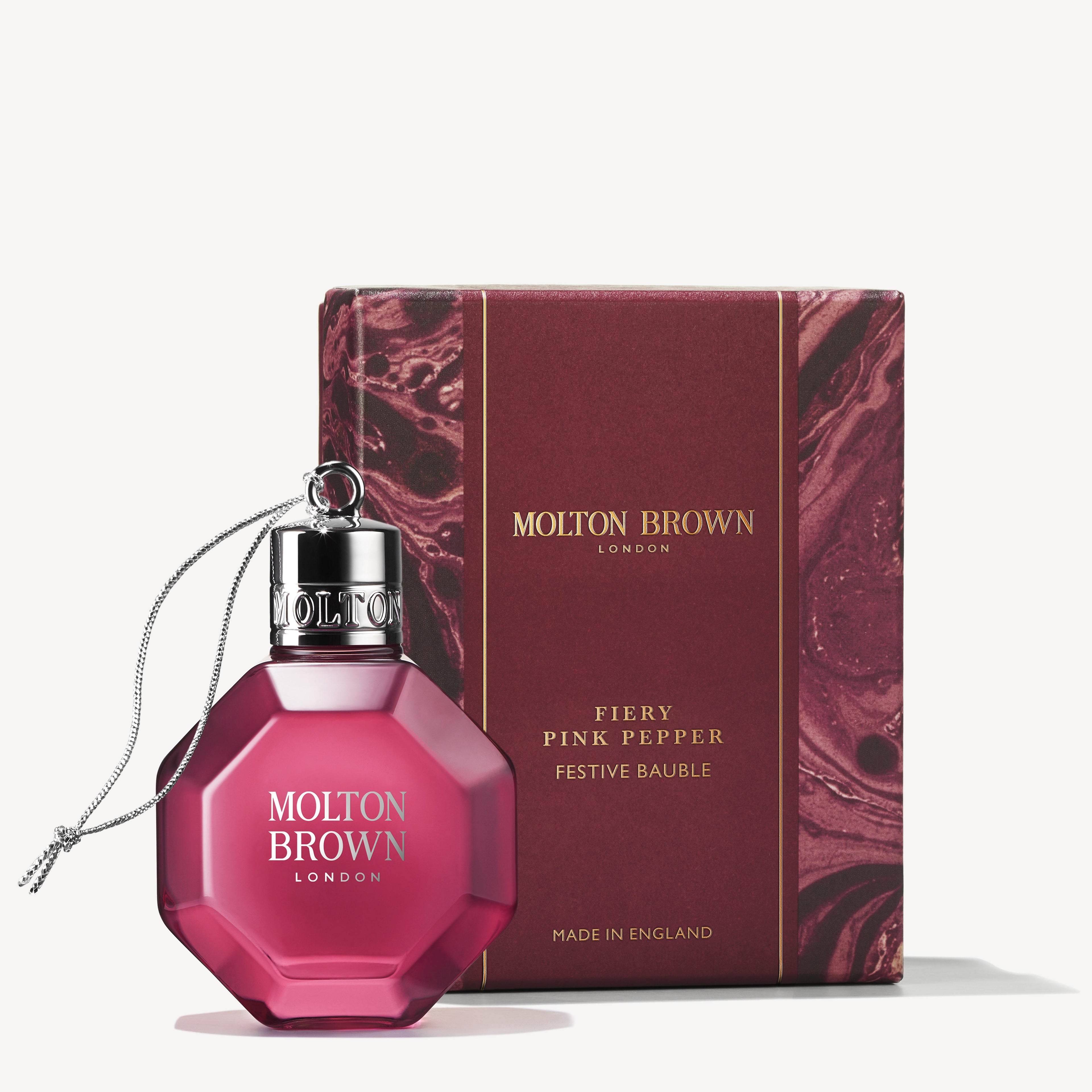 A hexagonal pink perfume bottle with a shiny silver cap is adorned with a festive string. Next to it rests a matching maroon box featuring gold text: MoltonBrown Cyprus, Fiery Pink Pepper Festive Bauble, Made in England.