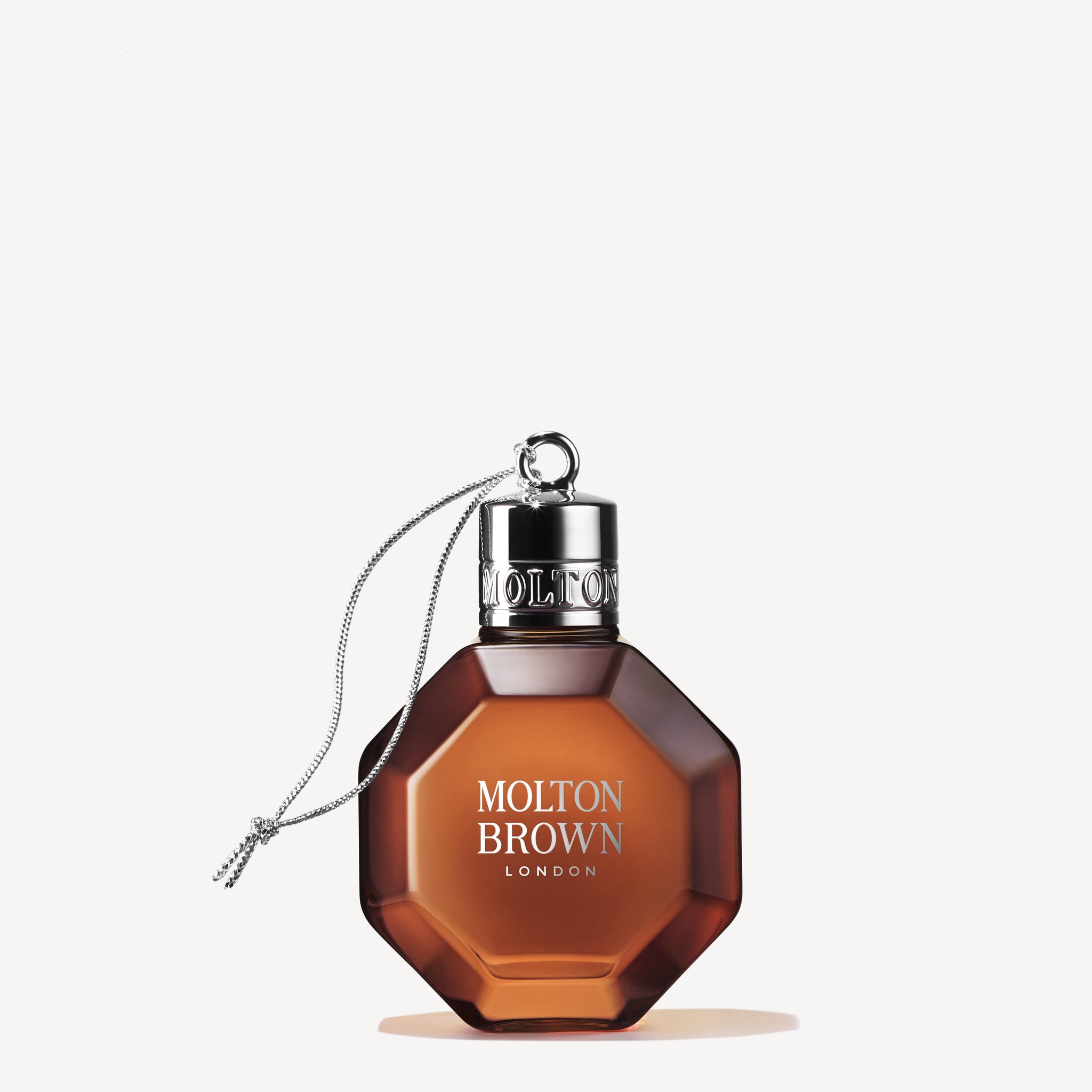 A geometric amber-colored perfume bottle labeled MoltonBrown Cyprus, featuring the Re-charge Black Pepper Festive Bauble design, with a metallic cap and a silver chain. The presentation is reminiscent of the spicy coriander scent, set against a plain white background.