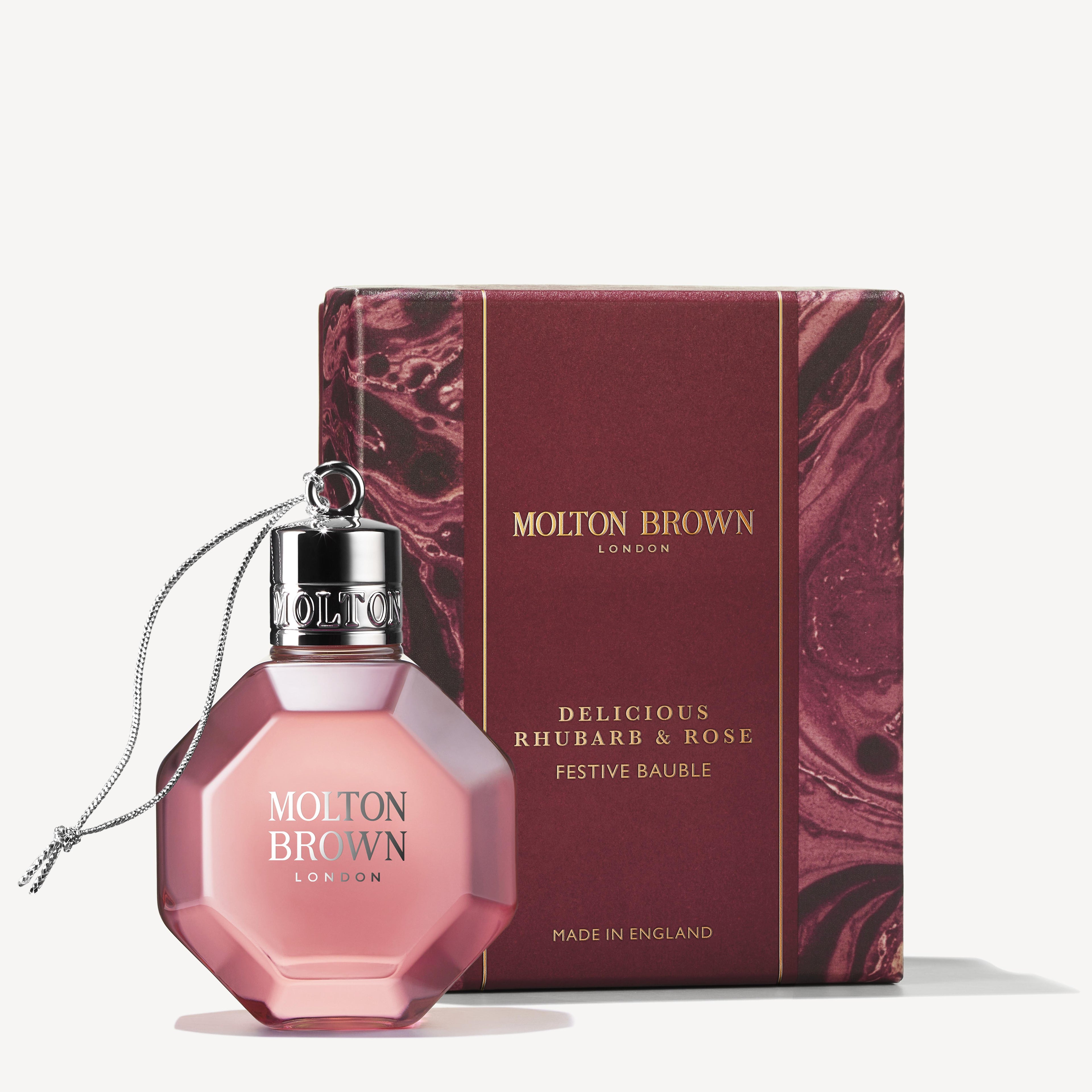 A pink, hexagonal MoltonBrown Cyprus perfume bottle labeled Delicious Rhubarb & Rose Festive Bauble, complete with a silver cap and string, sits beside its maroon box adorned with a marble pattern and branding. Infused with notes of grapefruit, this fragrance captures the charm of a festive bauble.