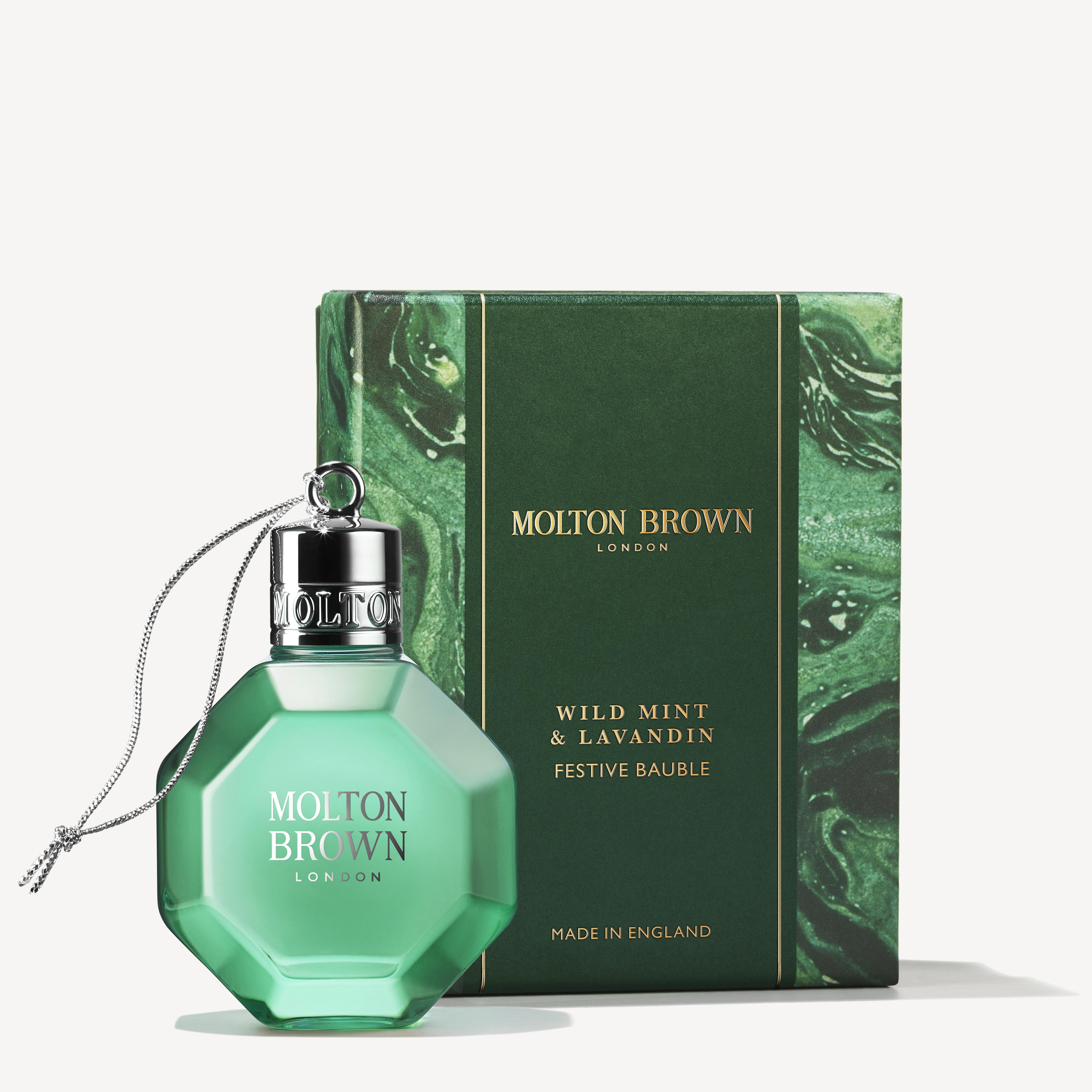 A festive bauble fragrance bottle from MoltonBrown Cyprus, in light green and featuring a silver cap and string, is displayed in front of a matching green box labeled Wild Mint & Lavandin Festive Bauble, with text noting it is proudly made in England.