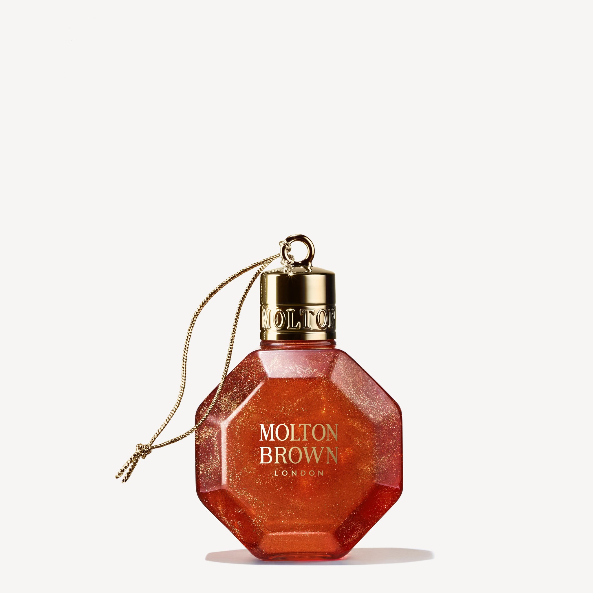 The Marvellous Mandarin & Spice Festive Bauble by MoltonBrown Cyprus is a hexagonal, translucent orange bottle designed to resemble a Christmas bauble. It features a gold cap and includes a hanging cord. Set against a plain, light background, this glistening bottle captures the refreshing charm of winter citrus.