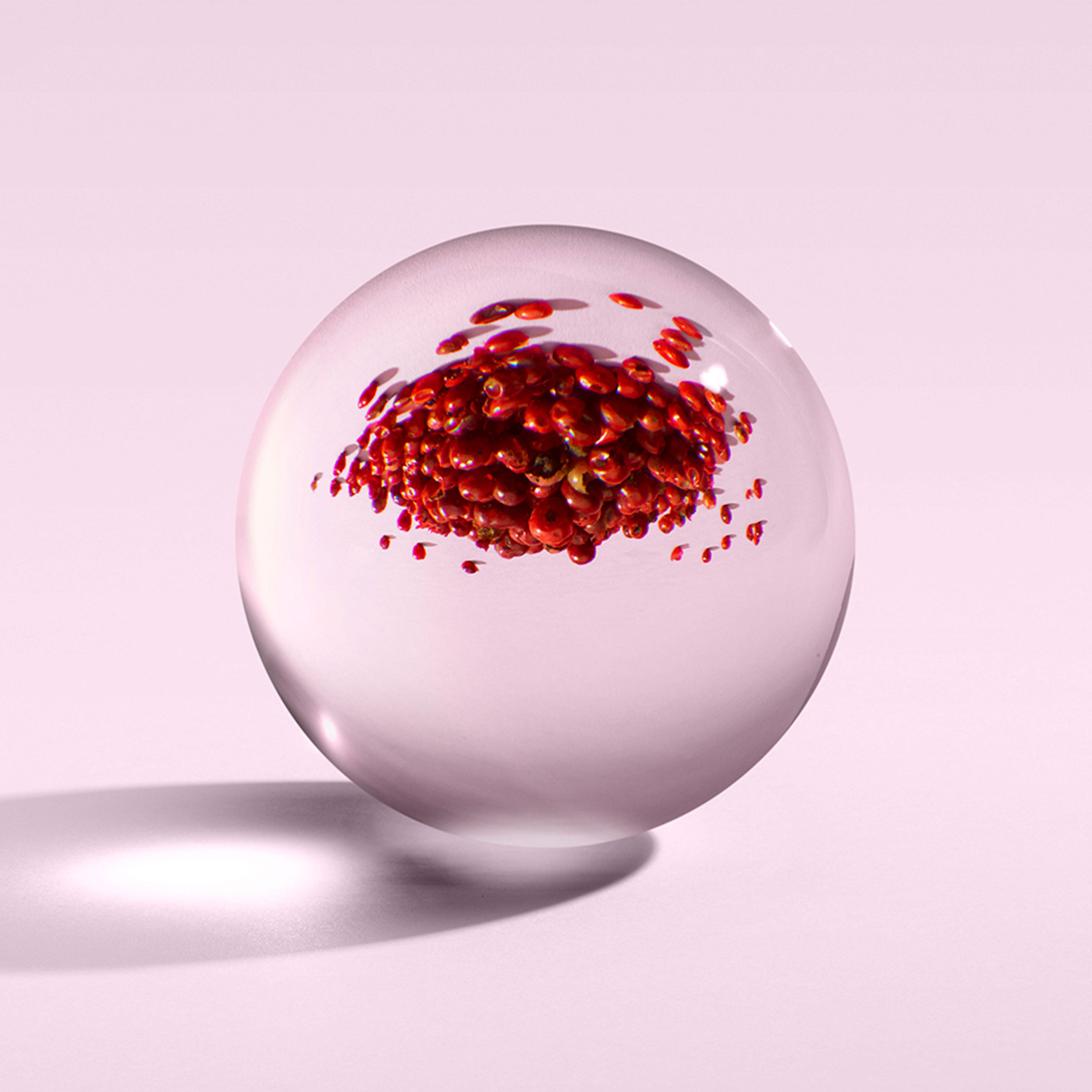 A clear glass sphere placed on a pink background encases a cluster of red berries, their reflection shimmering like the appearance of Fiery Pink Pepper Bath & Shower Gel from MoltonBrown Cyprus on the sphere's surface. Shadows stretch to the left.