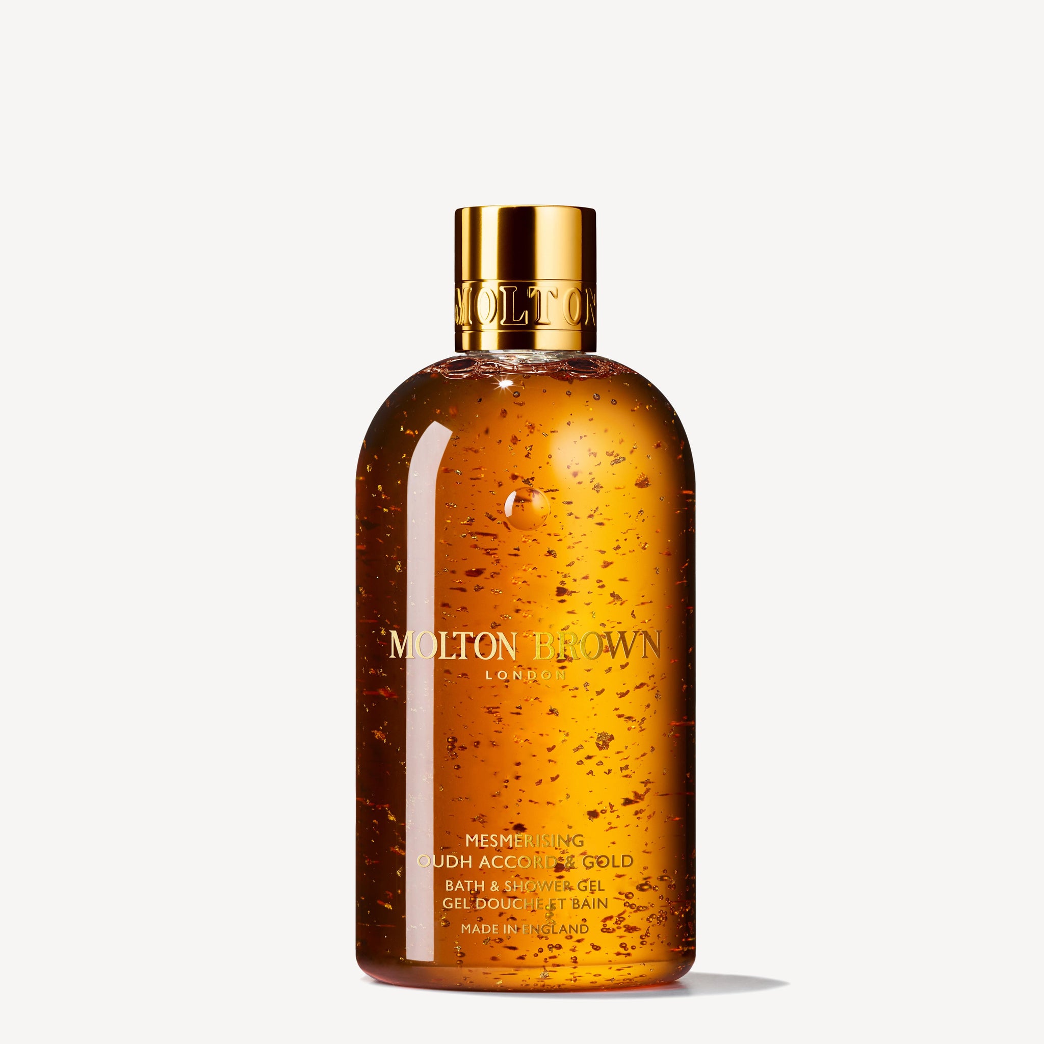 A bottle of MoltonBrown Cyprus Mesmerising Oudh Accord & Gold Bath & Shower Gel 300ml features a transparent design embellished with golden flakes that evoke the essence of a spiced forest, and is completed with a radiant golden cap, all set against a plain background.