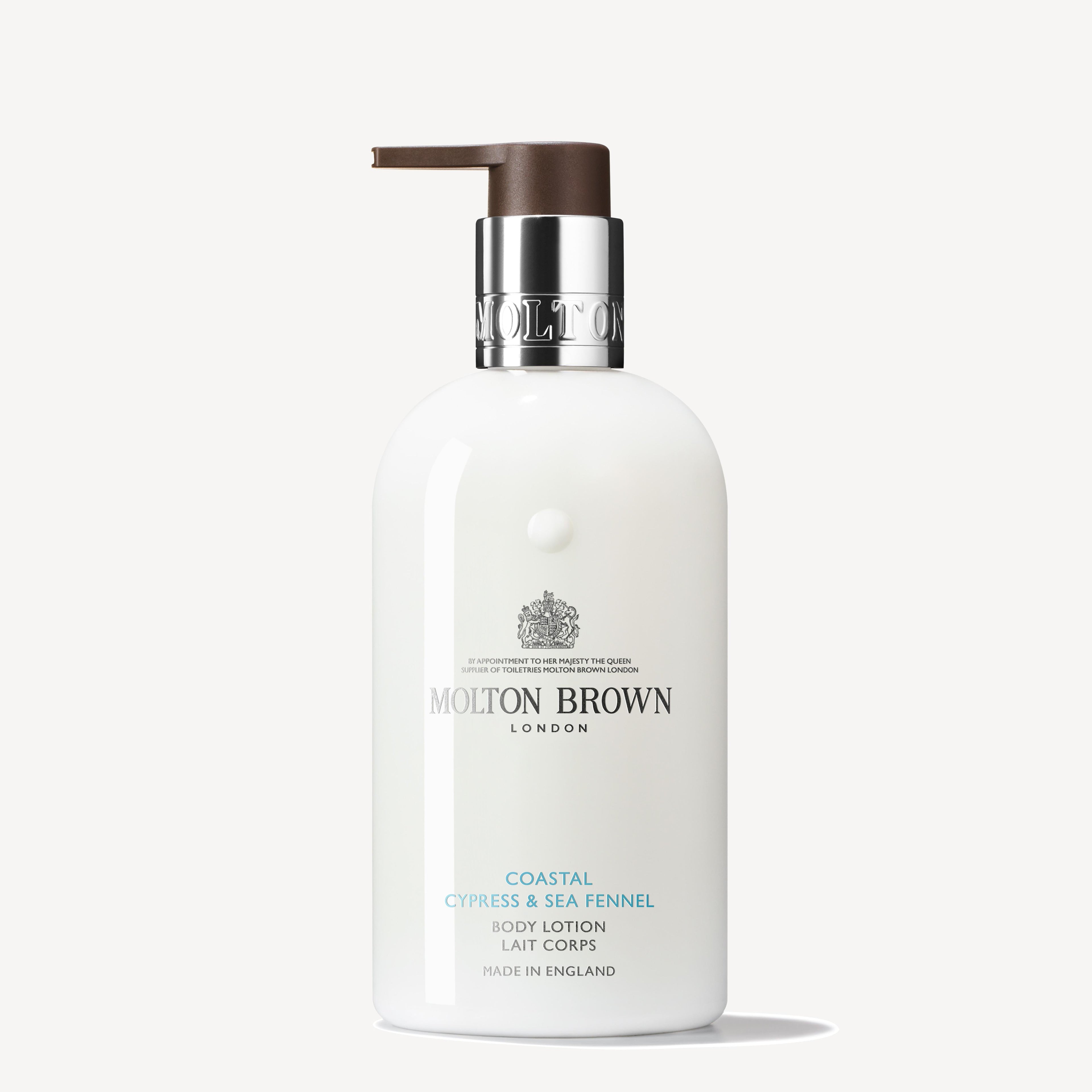 A 300ml bottle of MoltonBrown Cyprus Coastal Cypress & Sea Fennel Body Lotion, featuring a stylish white design with a silver and brown pump dispenser, elegantly displays the brand's logo and text in silver and blue while hydrating the skin beautifully.