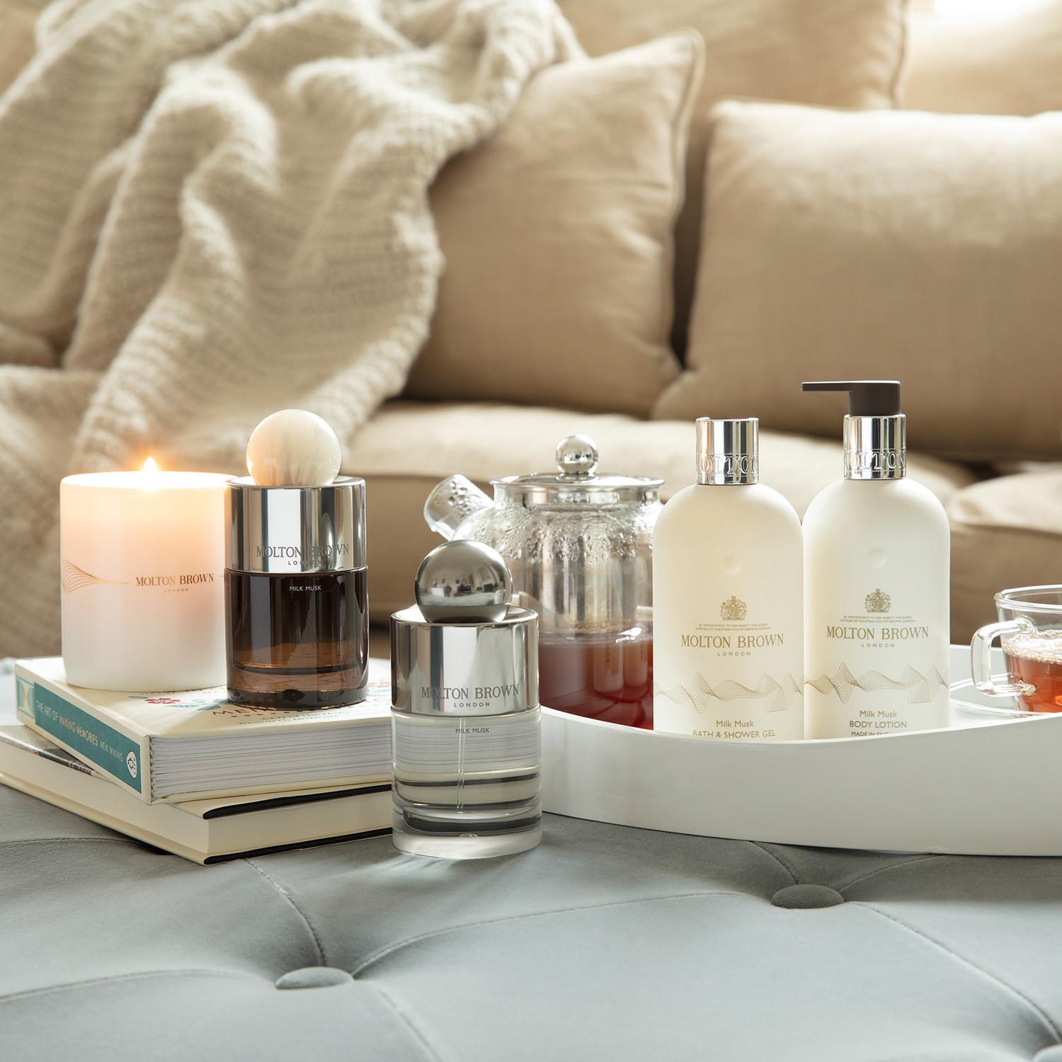 A cozy setting features the Milk Musk Body Lotion 300ml by MoltonBrown Cyprus on a tray, accompanied by a lit candle and teapot. In the background, books and a soft beige blanket enhance the warm ambiance filled with creamy musk notes.