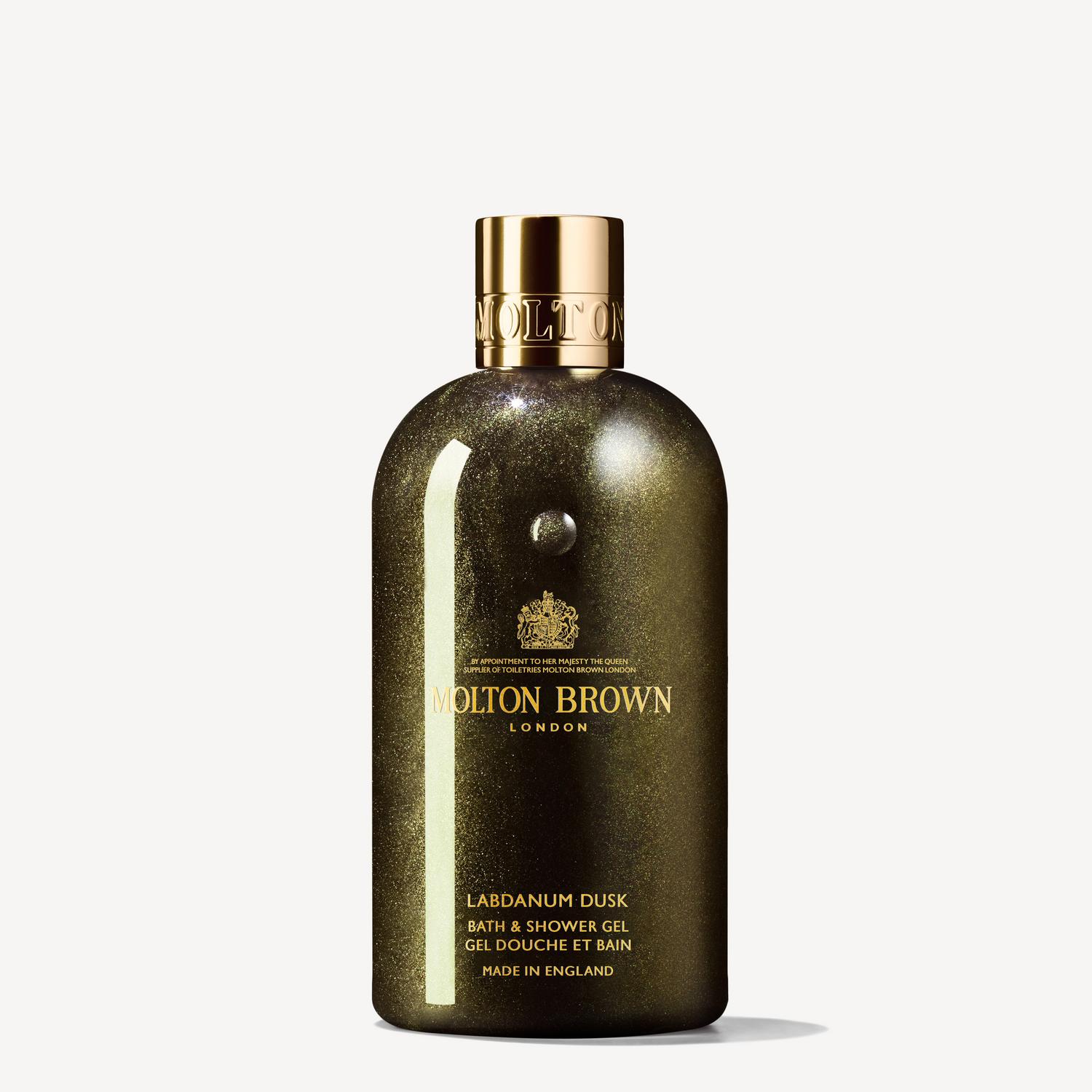 The image showcases a Labdanum Dusk Bath & Shower Gel 300ml bottle from MoltonBrown Cyprus, featuring its glittery dark green packaging accented by a gold cap and lettering. This luxurious presentation invites you to indulge in an ambery-woody fragrance with hints of opulent oudh. Proudly displayed is the note "Made in England.