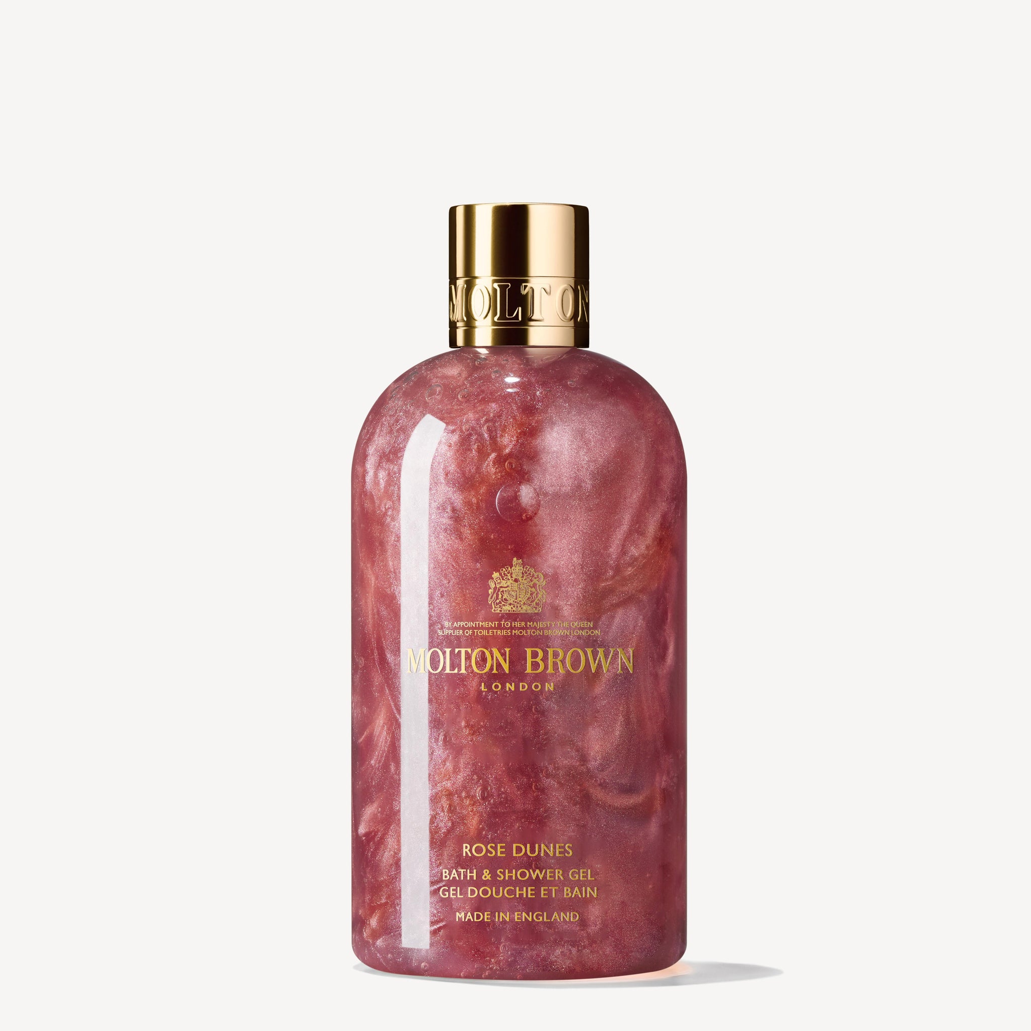 The Rose Dunes Bath & Shower Gel 300ml from MoltonBrown Cyprus features a pink, marbled liquid infused with patchouli Sulawesi, and comes with a gold cap and label adorned with elegant branding and text.