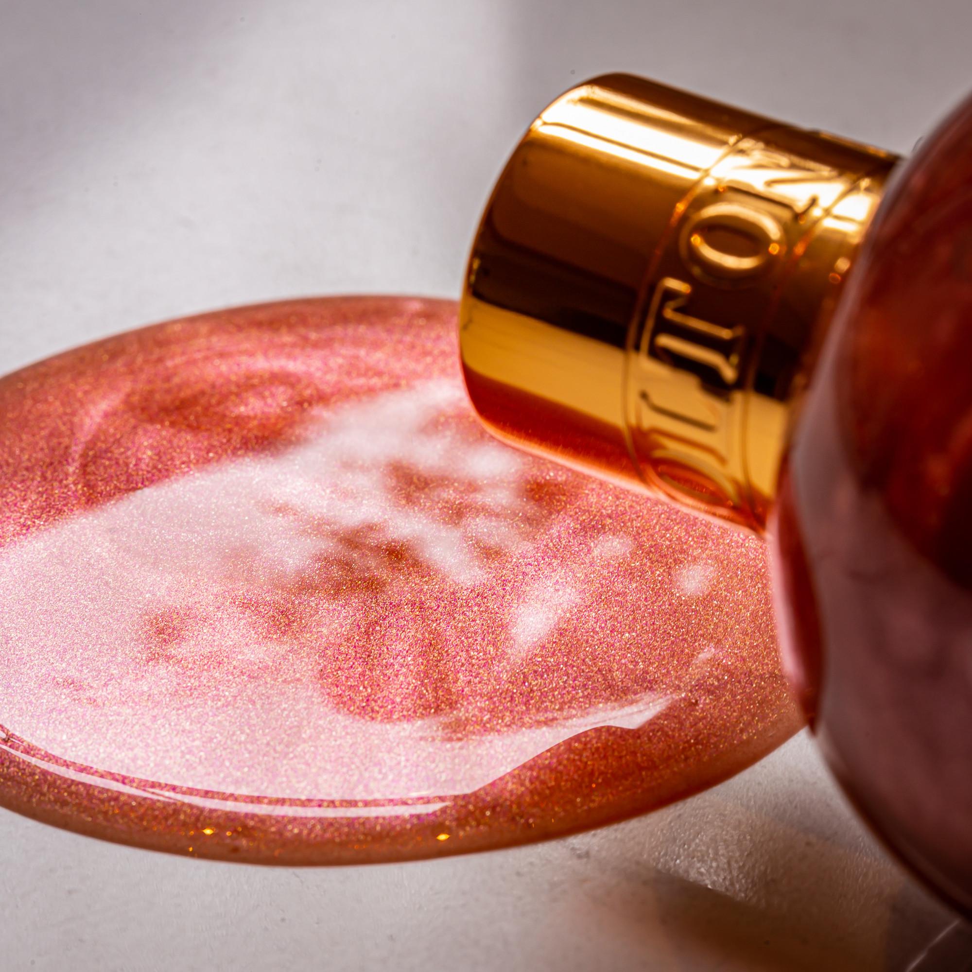 A metallic bottle labeled Rose Dunes Bath & Shower Gel by MoltonBrown Cyprus, featuring a gold cap, rests on its side as it pours a shimmering pink liquid onto a flat surface. This ambery body wash sparkles under the light, forming a glossy pool. The close-up scene is vibrant and captivating.