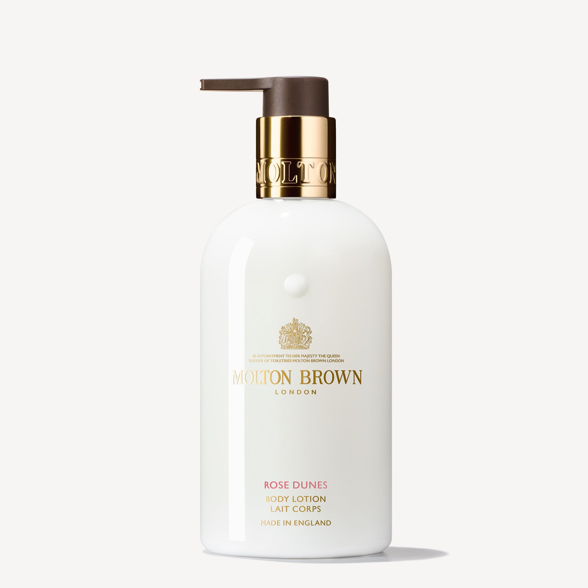 A white bottle of MoltonBrown Cyprus's Rose Dunes Body Lotion 300ml features a gold pump dispenser and offers an ambery-rose scent. The label displays the brand's logo and product details in gold text against a plain off-white background, enhancing its elegance and promise of hydration.