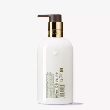 A white bottle with a pump dispenser, highlighted by gold accents near the top, stands against a minimalistic background. The text and symbols on the bottle reveal it as MoltonBrown Cyprus's Merry Berries & Mimosa Body Lotion, hinting at its winter skin formula infused with red berries.