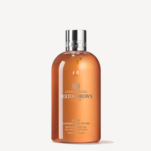 A 300ml bottle of Sunlit Clementine & Vetiver Bath & Shower Gel by MoltonBrown Cyprus. This clear bottle displays an orange liquid and is topped with a silver cap. The brand logo and product details are elegantly printed in white on the front, offering a refreshing citrus body wash experience.