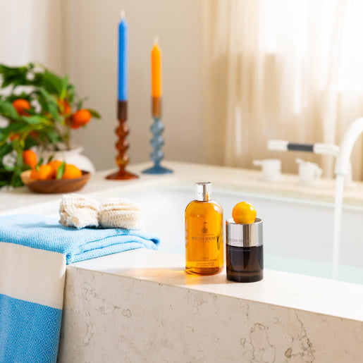 A bathroom setting with two bottles on a bathtub ledge: one amber bottle of MoltonBrown Cyprus Sunlit Clementine & Vetiver Bath & Shower Gel 300ml, offering a refreshing citrus aroma, and another dark bottle, rich with lemon. A blue towel and bath accessories are nearby. In the background, two lit candles and a bowl of clementines evoke tranquility.