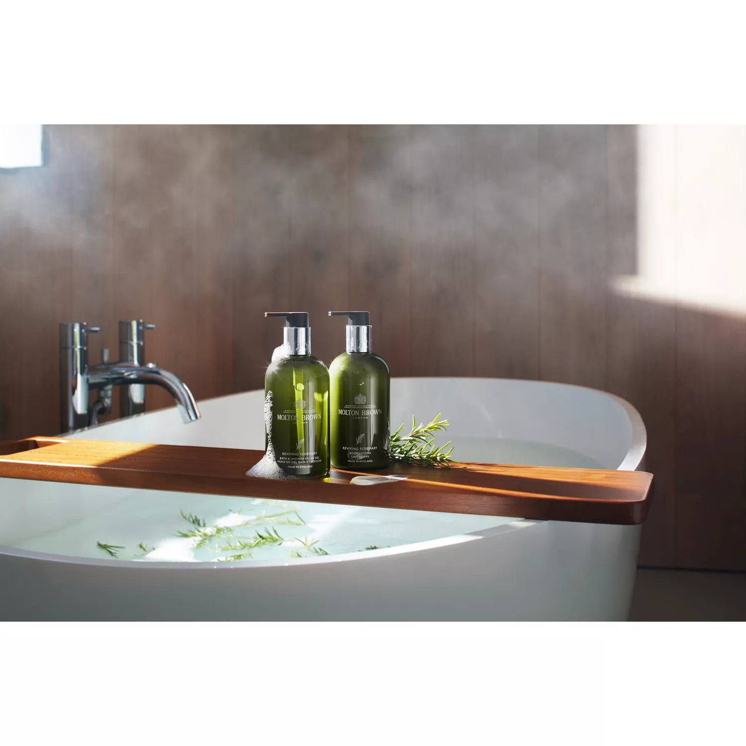 Two green MoltonBrown Cyprus bottles with pump dispensers, each holding Reviving Rosemary Bath & Shower Oil-In-Gel 300ml, rest on a wooden tray over a steaming bathtub. Sprigs of greenery add a natural touch; in the background, the modern faucet gleams. A luxurious milky lather awaits.