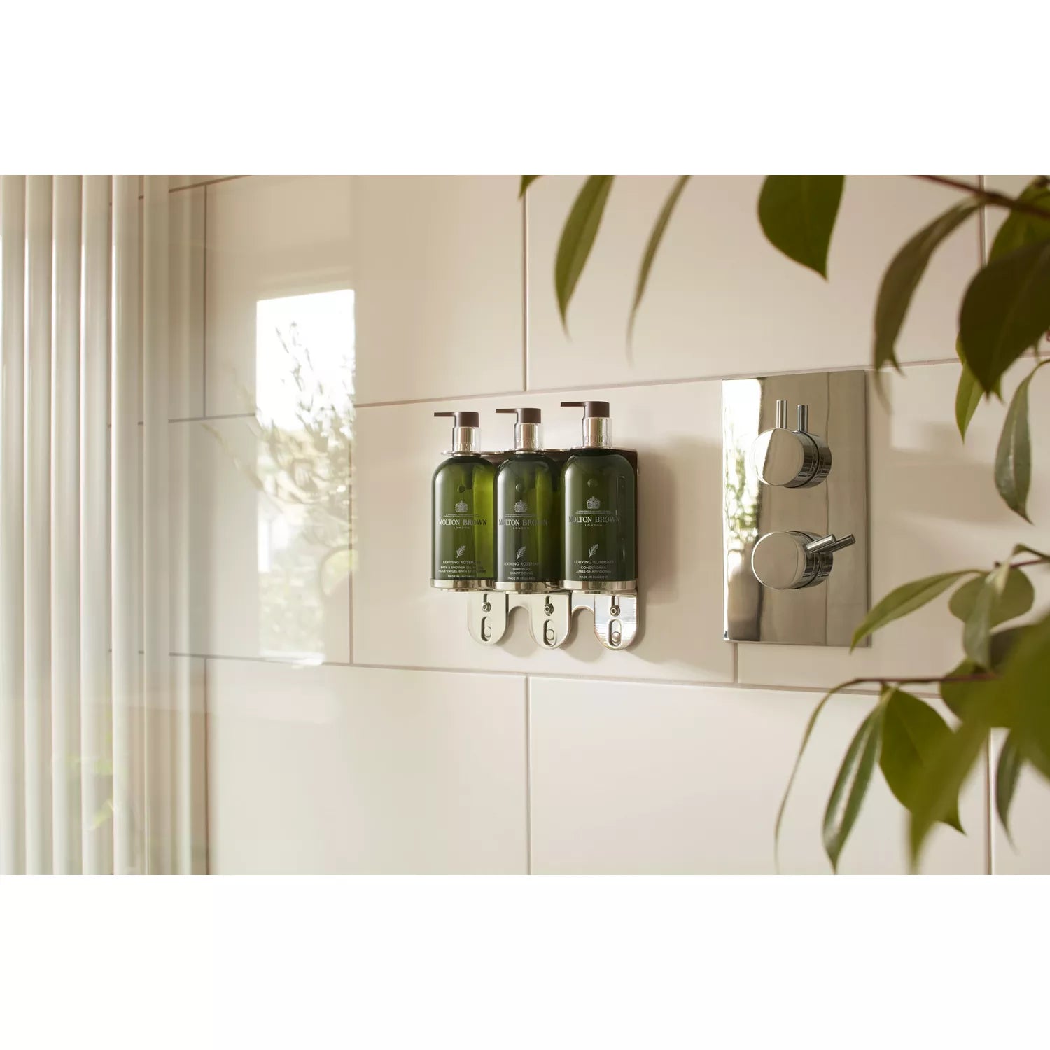 Wall-mounted shower dispensers hold three MoltonBrown Cyprus Reviving Rosemary Bath & Shower Oil-In-Gel 300ml bottles in a cream-tiled bathroom, adjacent to a chrome shower control. Natural light and nearby plant leaves enhance the serene atmosphere with reflections.