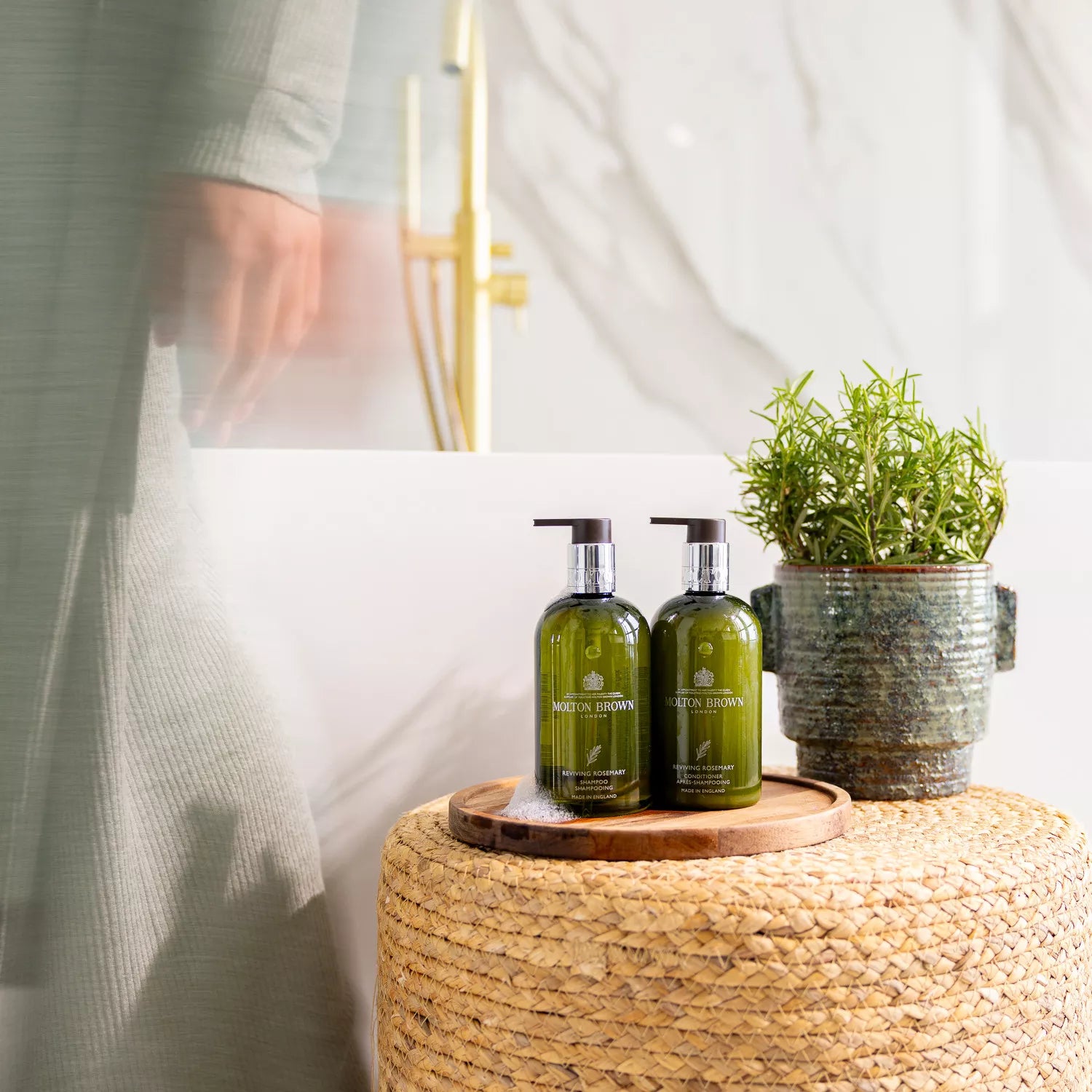 Two bottles of Molton Brown Cyprus Reviving Rosemary Shampoo, each 300ml with pump dispensers, sit on a woven surface by a potted plant. A person in a light robe is blurred in the background of the marble-walled bathroom, suggesting motion and promising healthier hair with every wash.