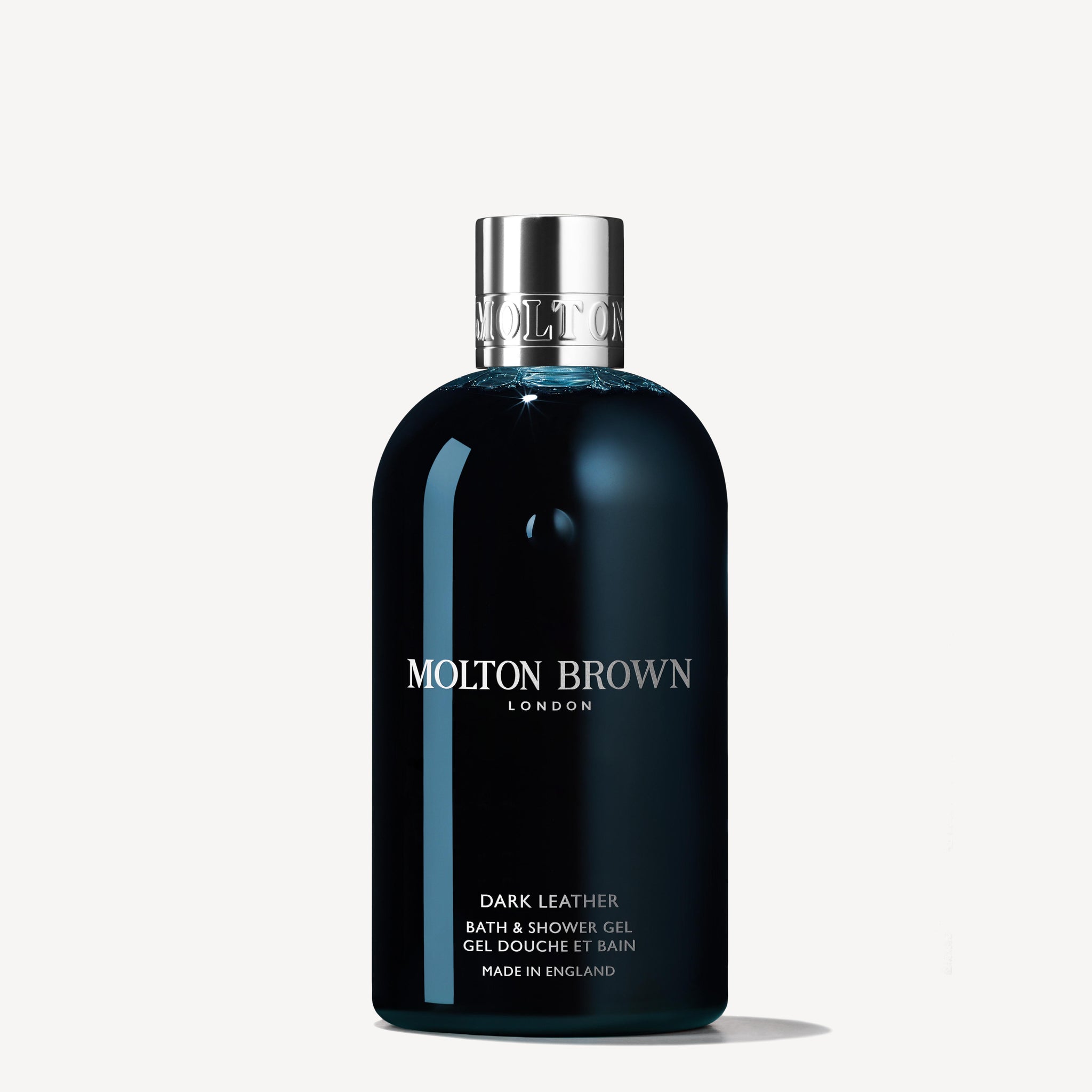 A 300ml bottle of MoltonBrown Cyprus Dark Leather Bath & Shower Gel, featuring a chrome cap, exudes sophistication. Its black label with white text prominently displays the brand's logo and product details, while notes of aged leather and a subtle tobacco accord enhance this luxurious experience against an off-white backdrop.