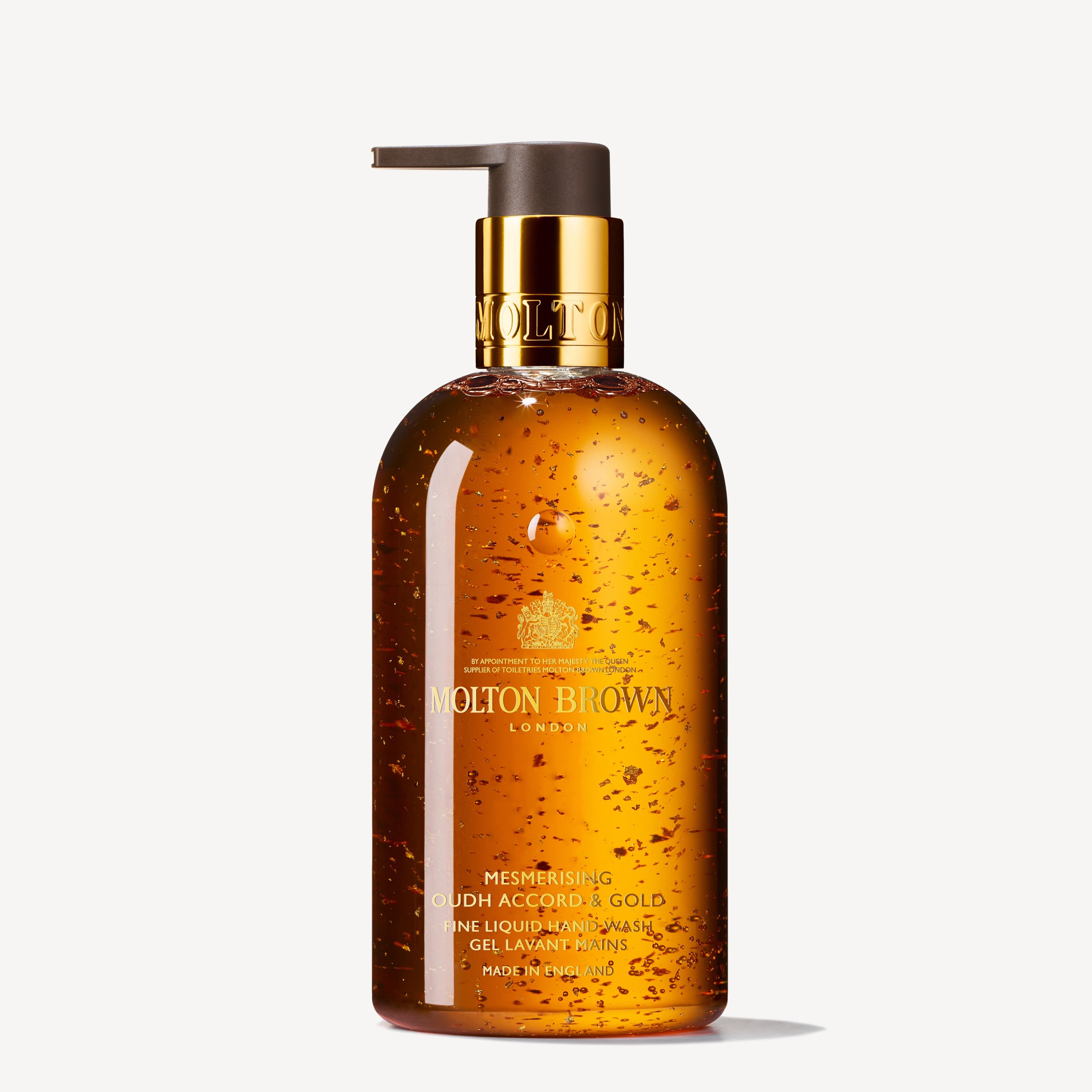 A 300ml bottle of MoltonBrown Cyprus Mesmerising Oudh Accord & Gold Fine Liquid Hand Wash, equipped with a pump dispenser. The elegant golden-brown design showcases 23.5-carat golden flakes shimmering inside, with the label prominently displaying the brand name and product title.