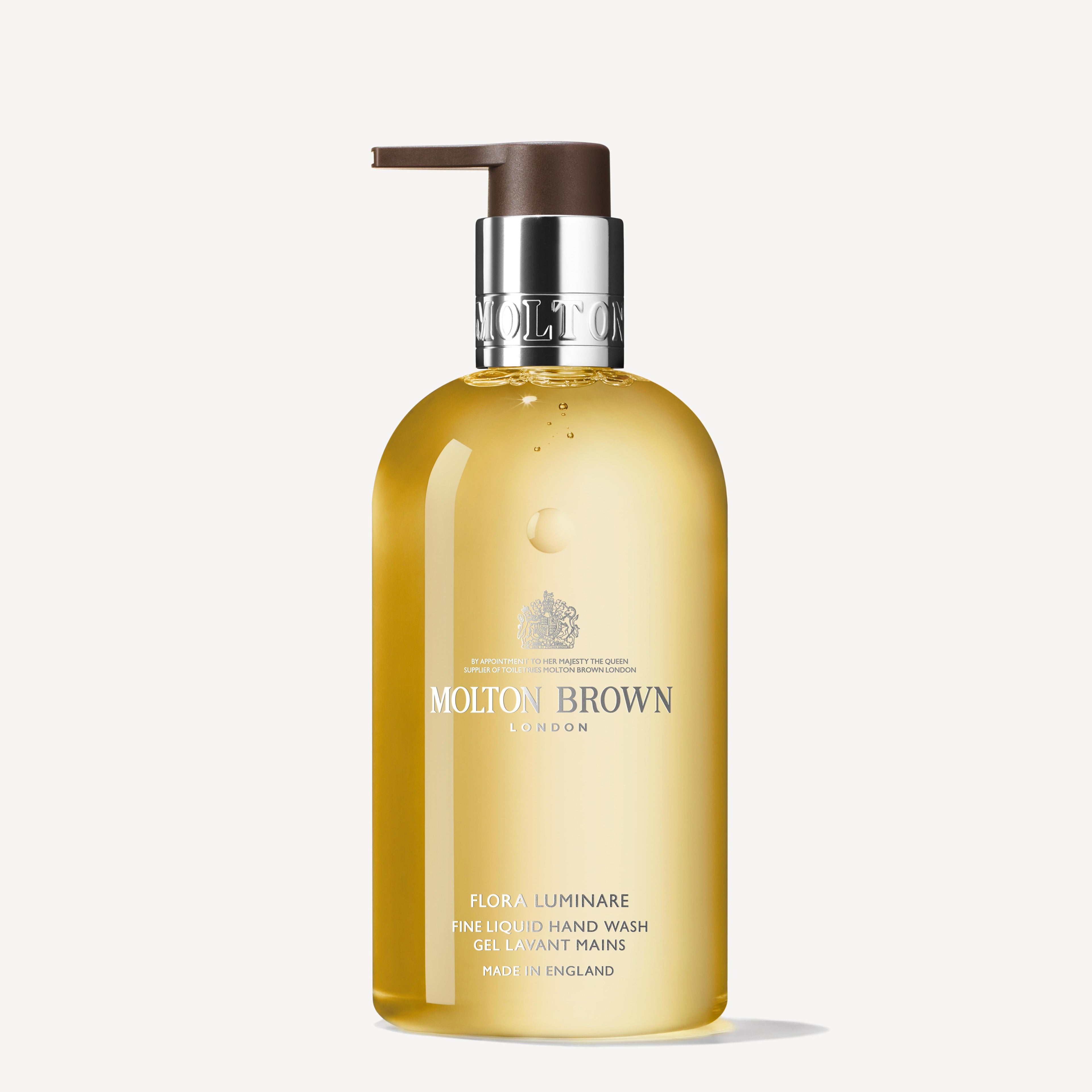 A 300ml bottle of MoltonBrown Cyprus's Flora Luminare Fine Liquid Hand Wash, highlighted with hints of orange blossom. Its transparent design showcases a yellow liquid and includes a silver label featuring text. The bottle features a brown pump dispenser and is crafted in England.