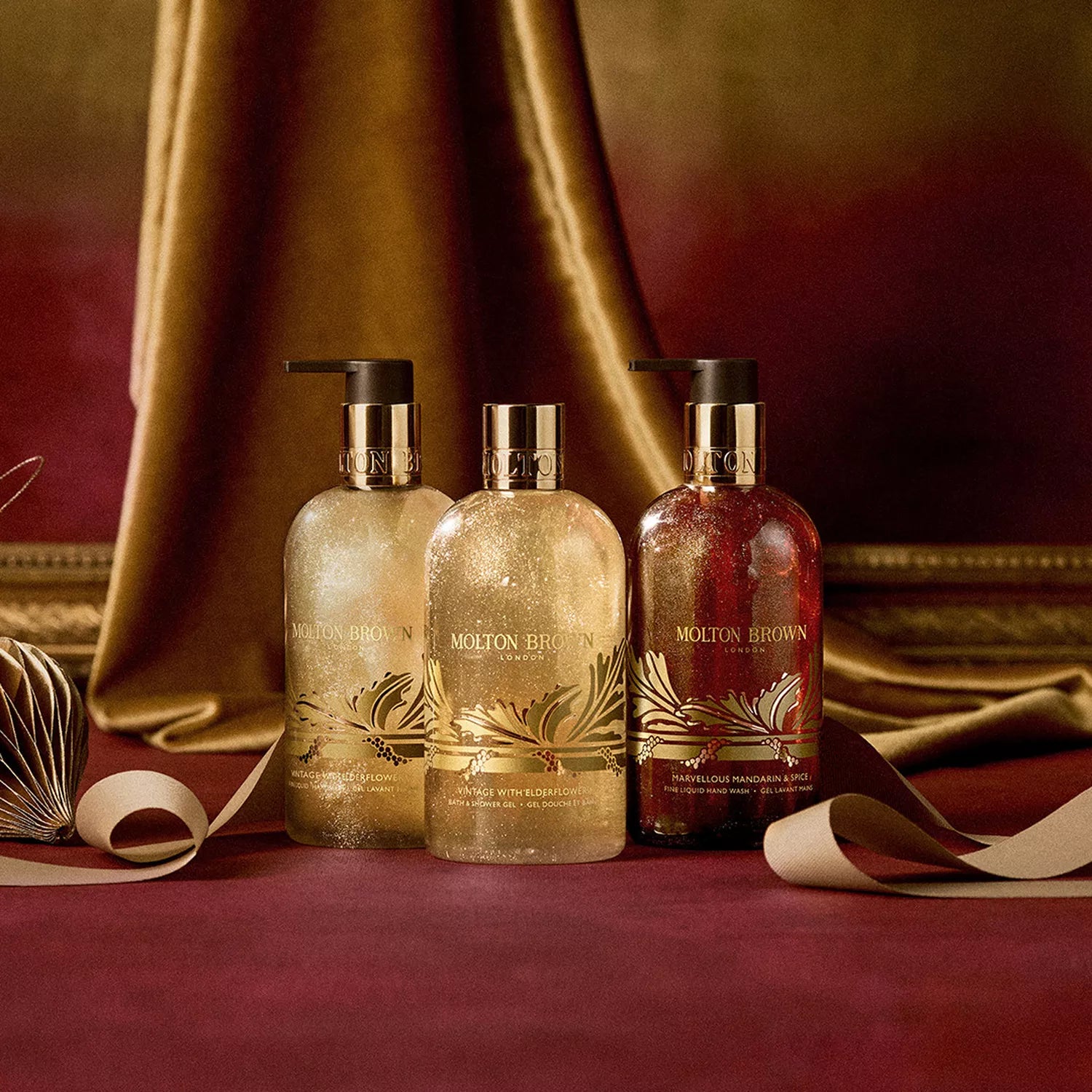 Three MoltonBrown Cyprus Vintage With Elderflower Fine Liquid Hand Wash 300ml bottles with gold and floral accents rest on red velvet, surrounded by a gold ribbon, creating a luxurious festive look with hints of floral and fruity scent.