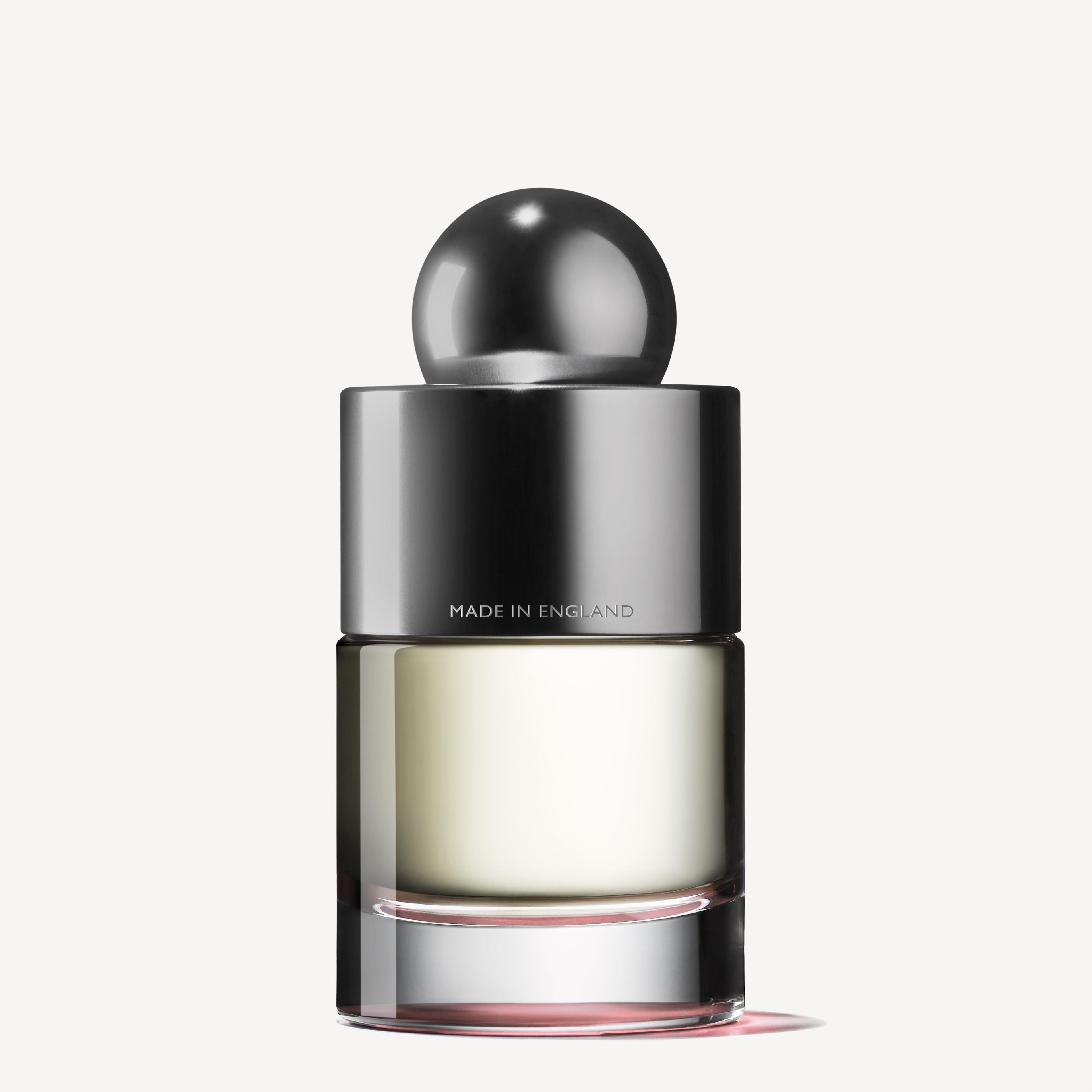 A sophisticated 100ml perfume bottle from MoltonBrown Cyprus features a smooth spherical black cap and a metallic silver collar, labeled MADE IN ENGLAND. This transparent bottle encases the Delicious Rhubarb & Rose Eau de Toilette, showcasing a light yellow liquid that suggests the scent of rhubarb leaf.