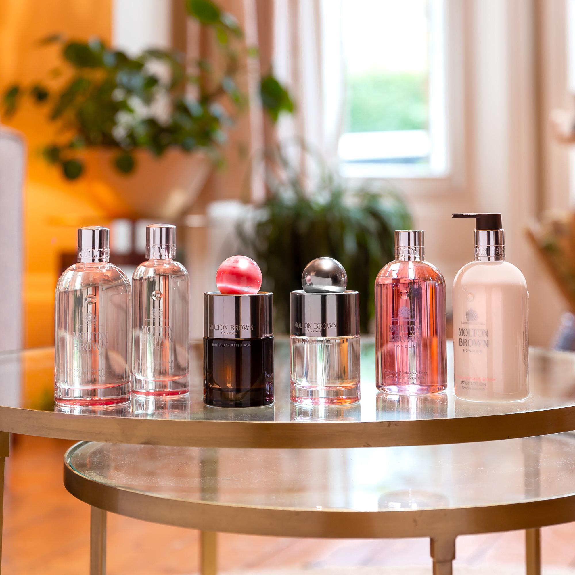 A collection of MoltonBrown Cyprus's Delicious Rhubarb & Rose Eau de Toilette 100ml perfumes and lotion adorns a glass table in a well-lit room. The bottles, diverse in size and color, exude elegance with notes of rhubarb leaf and grapefruit, while plants and windows accentuate the serene blurred background.