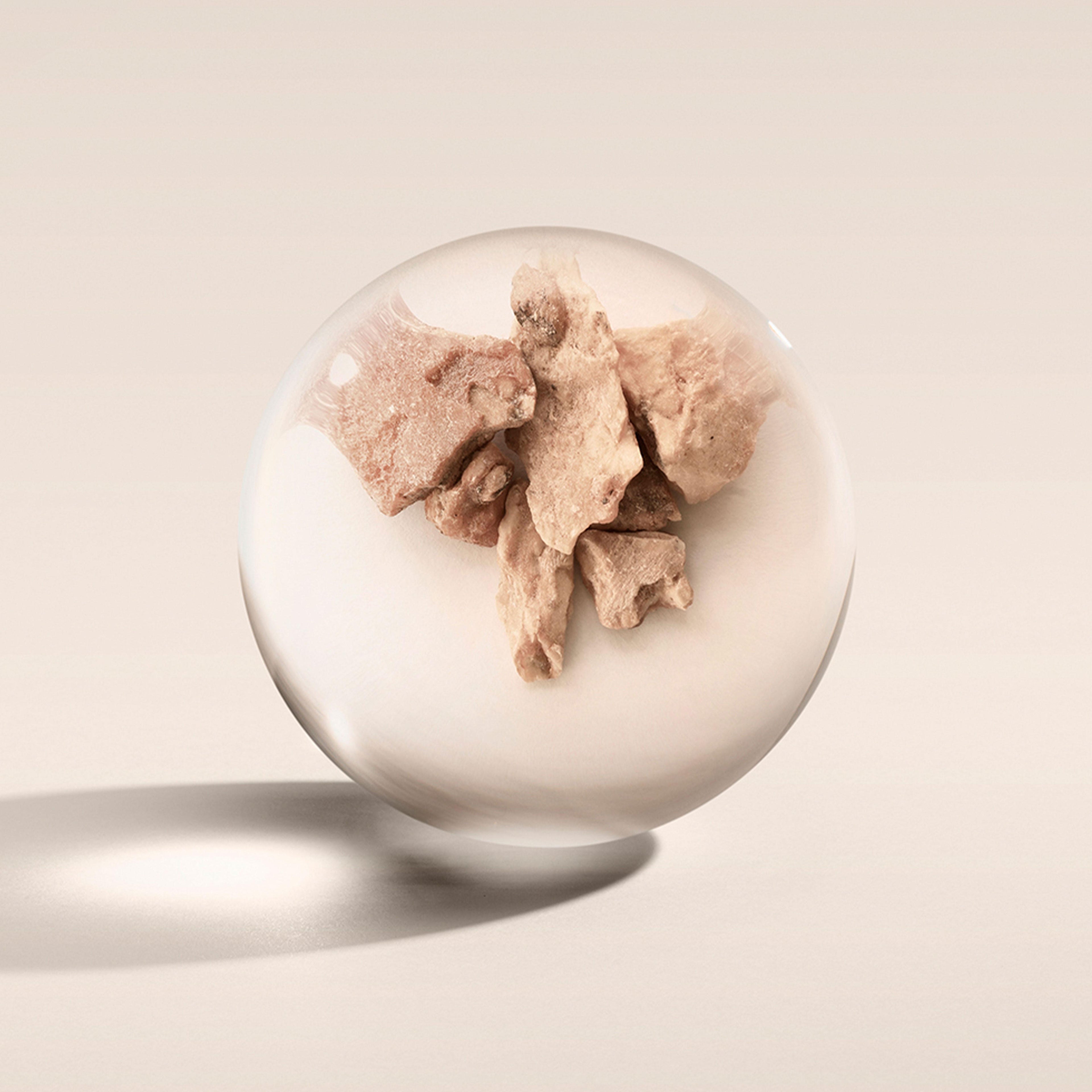 Inside a clear glass sphere, several irregularly shaped, brownish rocks or clay pieces are encased. The sphere casts a soft shadow on a light beige surface, creating an artistic composition that evokes the subtle allure of MoltonBrown Cyprus's Milk Musk Eau de Toilette 100ml with its floral-musky fragrance lingering in the air.