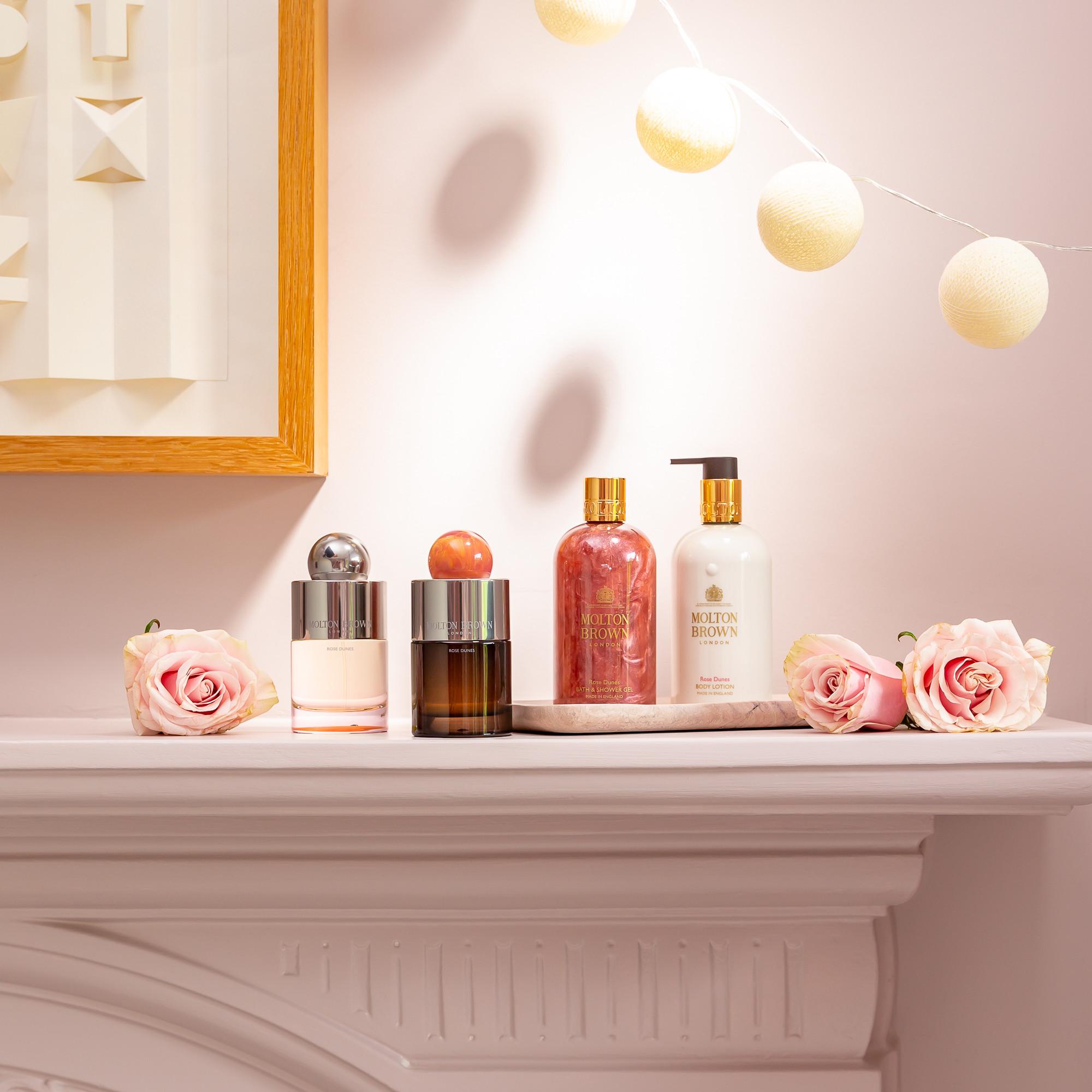 A white mantel showcases a collection of sophisticated Rose Dunes Eau de Toilette bottles from MoltonBrown Cyprus and lotion dispensers, accompanied by decorative items, including pink roses. The area is gently illuminated, with round, warm string lights hanging above to craft a cozy and inviting atmosphere.