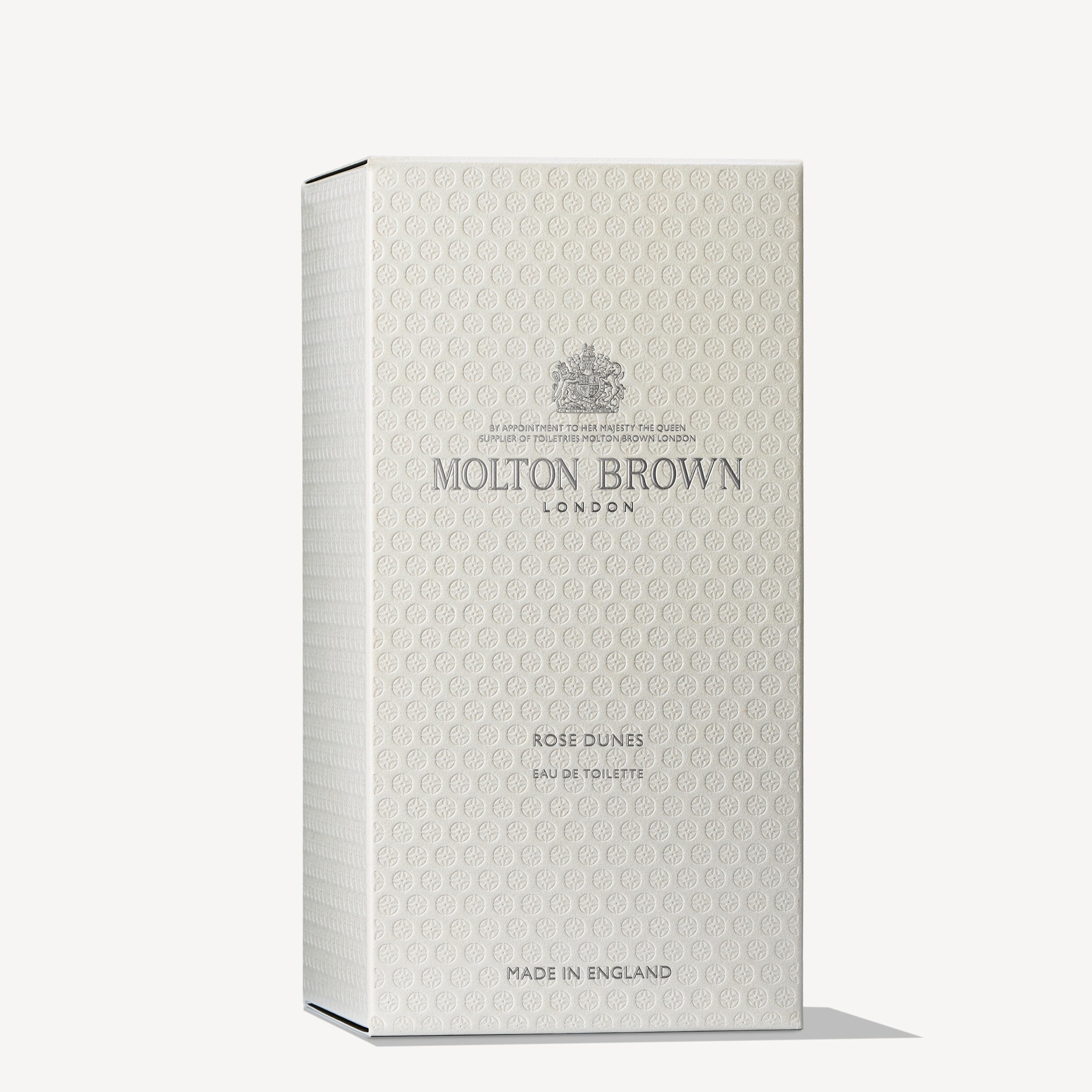 The cream-colored box of MoltonBrown Cyprus's Rose Dunes Eau de Toilette 100ml features an intricate embossed pattern and the brand's logo. With hints of cassis and patchouli Sulawesi, the fragrance exudes refined elegance. The text states "Made in England.