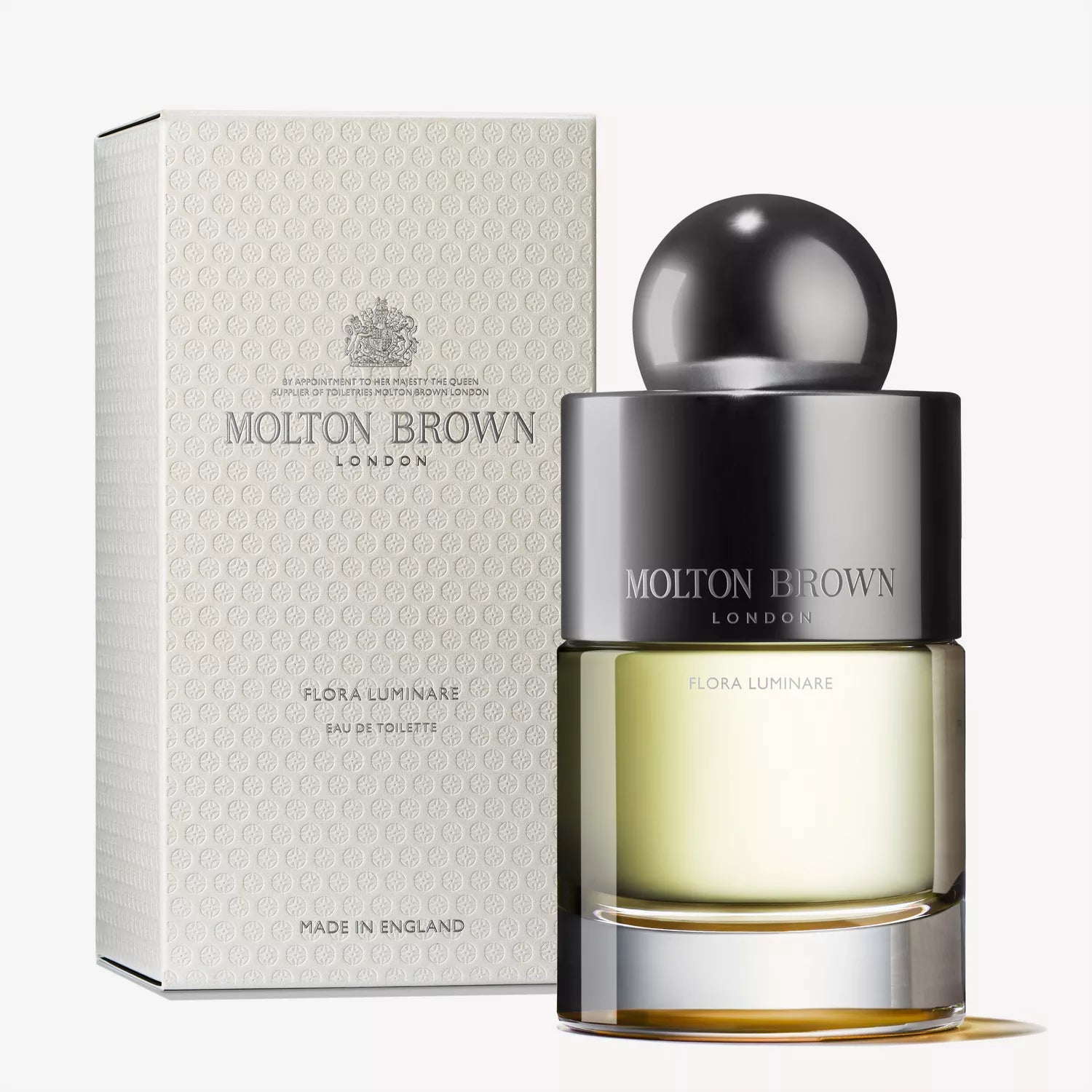 A Molton Brown Cyprus Flora Luminare Eau de Toilette 100ml bottle with a round cap is placed in front of its matching box, featuring an elegant design with branding in muted tones and hints of Polynesian frangipani.