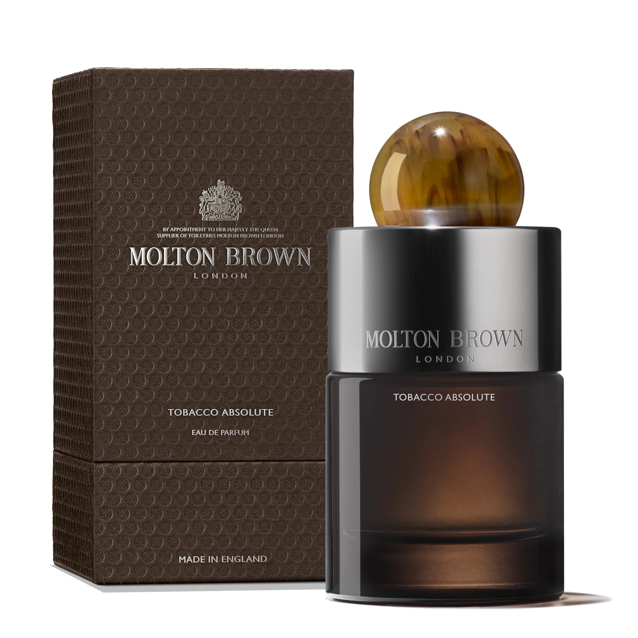 A bottle of MoltonBrown Cyprus Tobacco Absolute Eau de Parfum 100ml, offering a woody scent with smoky tobacco notes, is set before its textured brown packaging. The dark bottle is topped with a marbled brown spherical cap, and both items showcase elegant branding text.