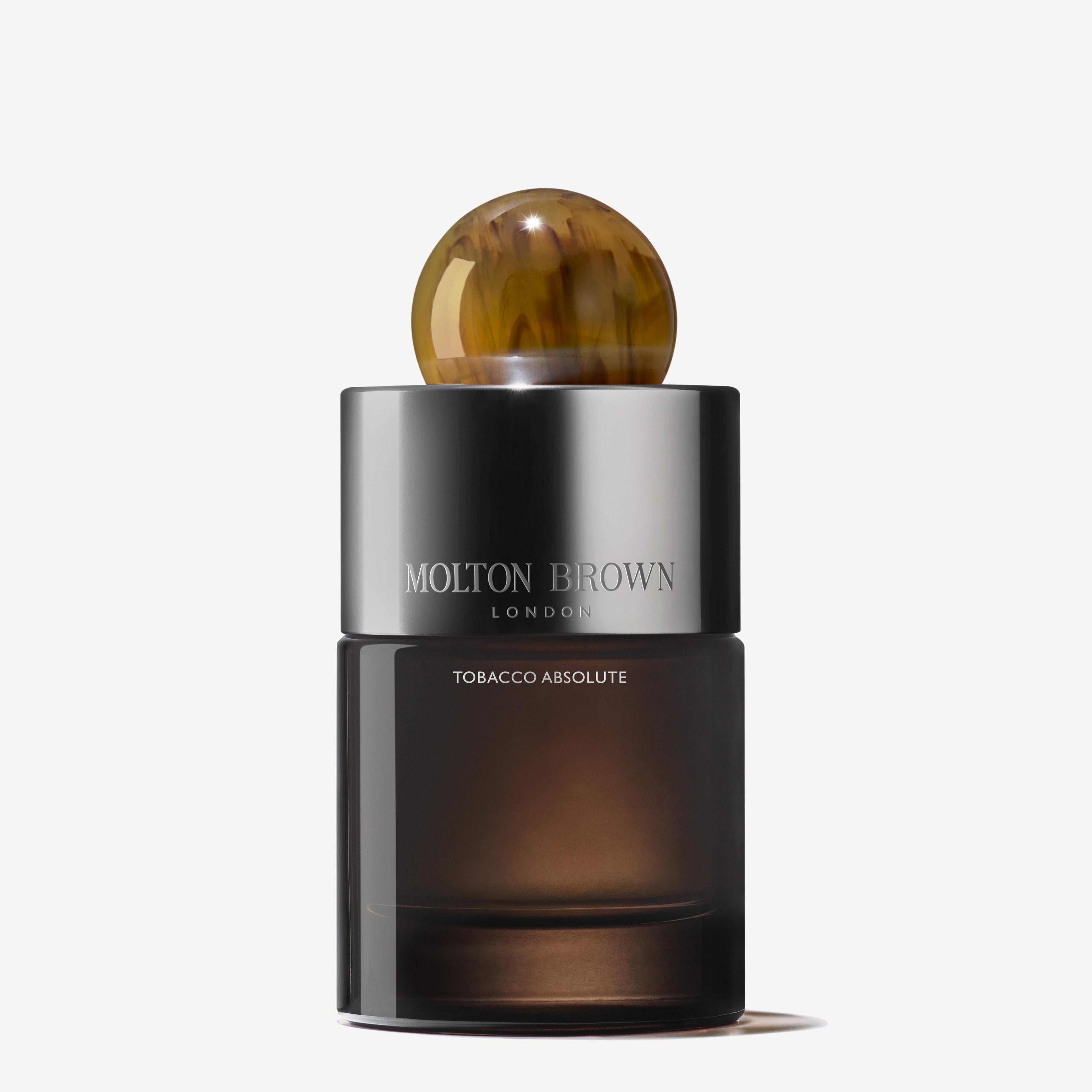 A dark, translucent cylindrical bottle of MoltonBrown Cyprus's Tobacco Absolute Eau de Parfum 100ml is shown. It boasts a shiny silver label and an amber-colored round cap, emitting a smoky tobacco allure. The plain, light-colored background accentuates its woody fragrance mystique.