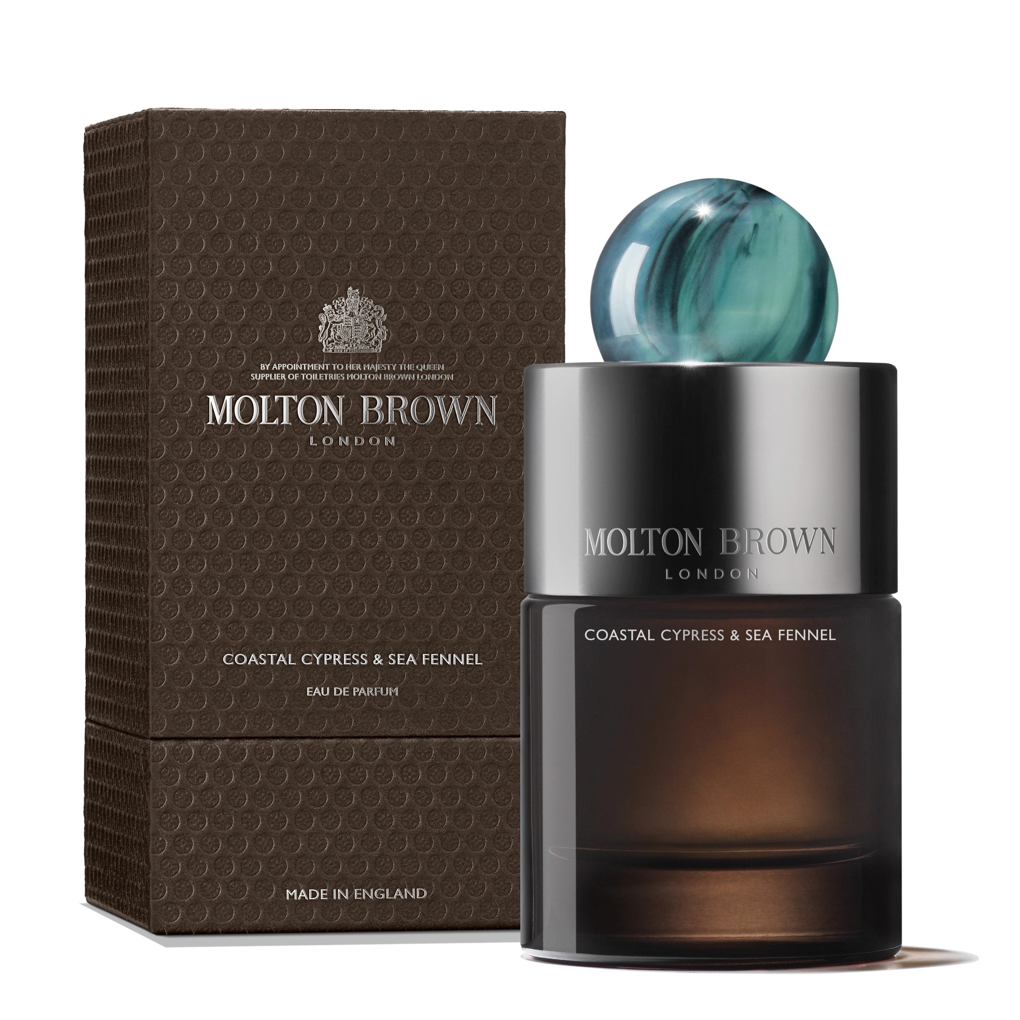 A MoltonBrown Cyprus perfume bottle, featuring a blue-green round cap, is positioned next to its textured dark brown packaging. The label indicates it is the Coastal Cypress & Sea Fennel Eau de Parfum 100ml, an ocean-inspired fragrance. Both the packaging and the bottle exhibit a sleek and elegant design.