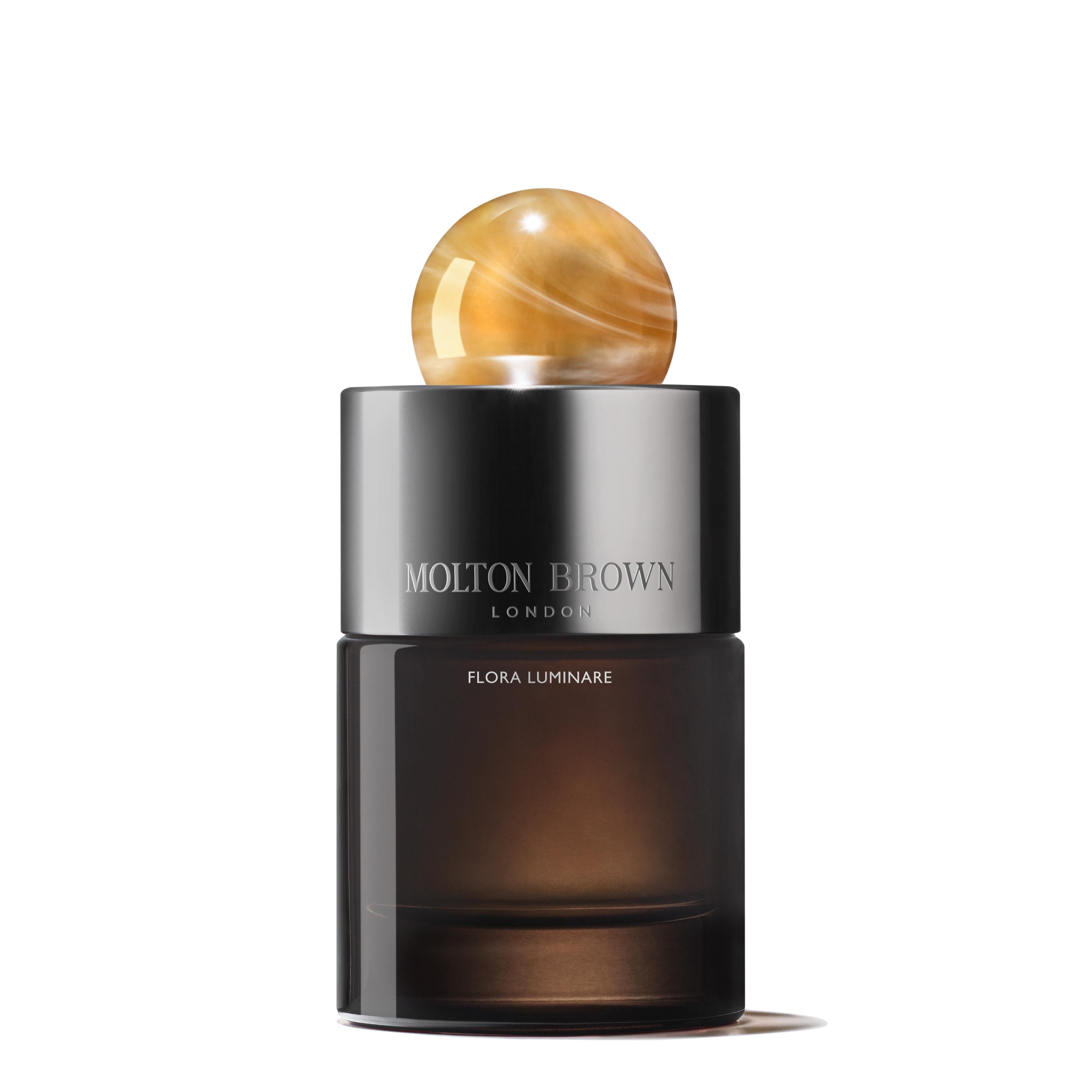 A sleek, dark bottle of Flora Luminare Eau de Parfum 100ml by MoltonBrown Cyprus, featuring a shiny amber-colored spherical cap. The refillable glass flacon exudes elegance with the bold text MOLTON BROWN CYPRUS printed on it. The design is minimalistic and sophisticated.