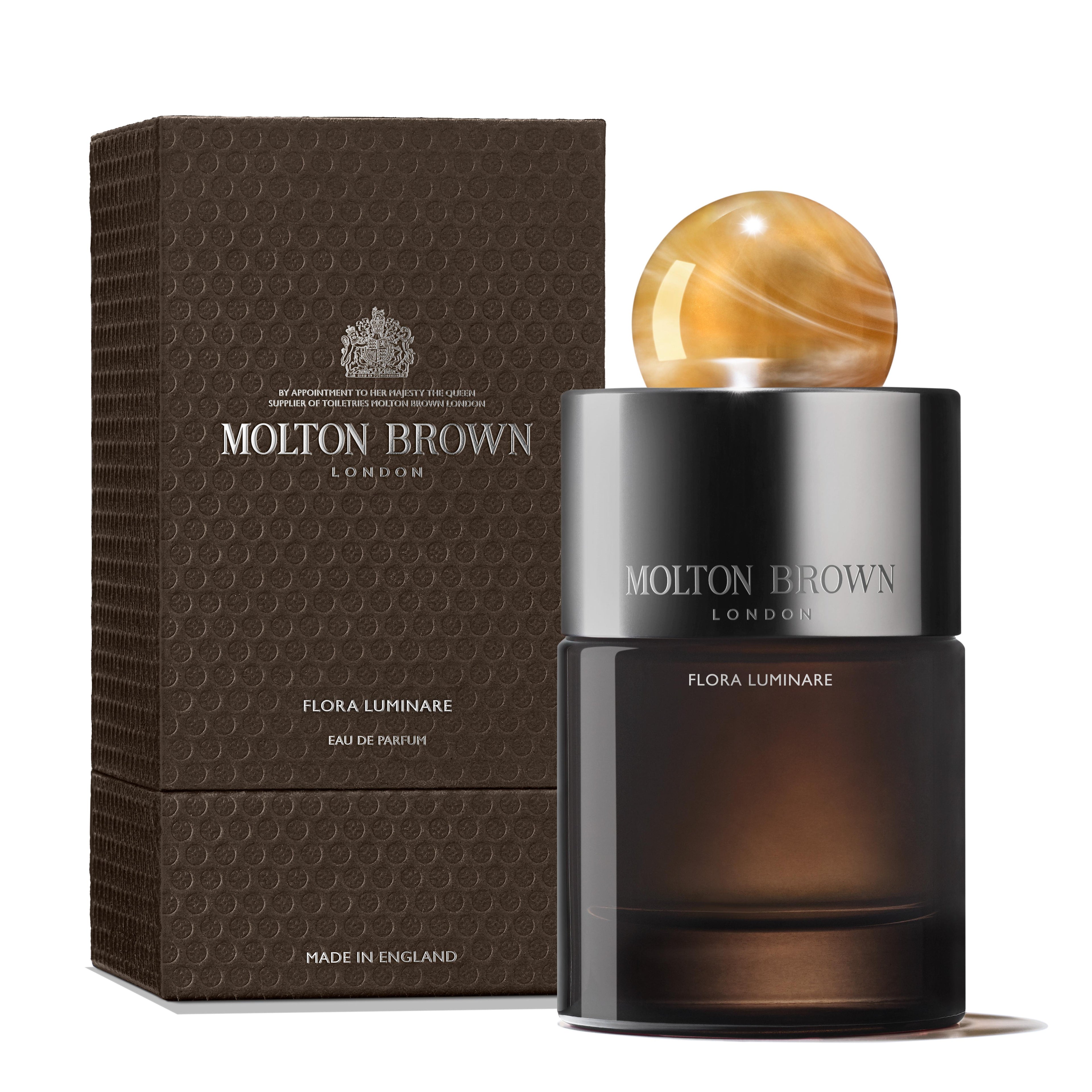 A luxurious MoltonBrown Cyprus Flora Luminare Eau de Parfum 100ml bottle, featuring a round marbled cap and a refillable glass flacon, stands beside a textured dark brown packaging box. The fragrance exudes hints of sandalwood. The text reads Molton Brown, London, Flora Luminare, Made in England.