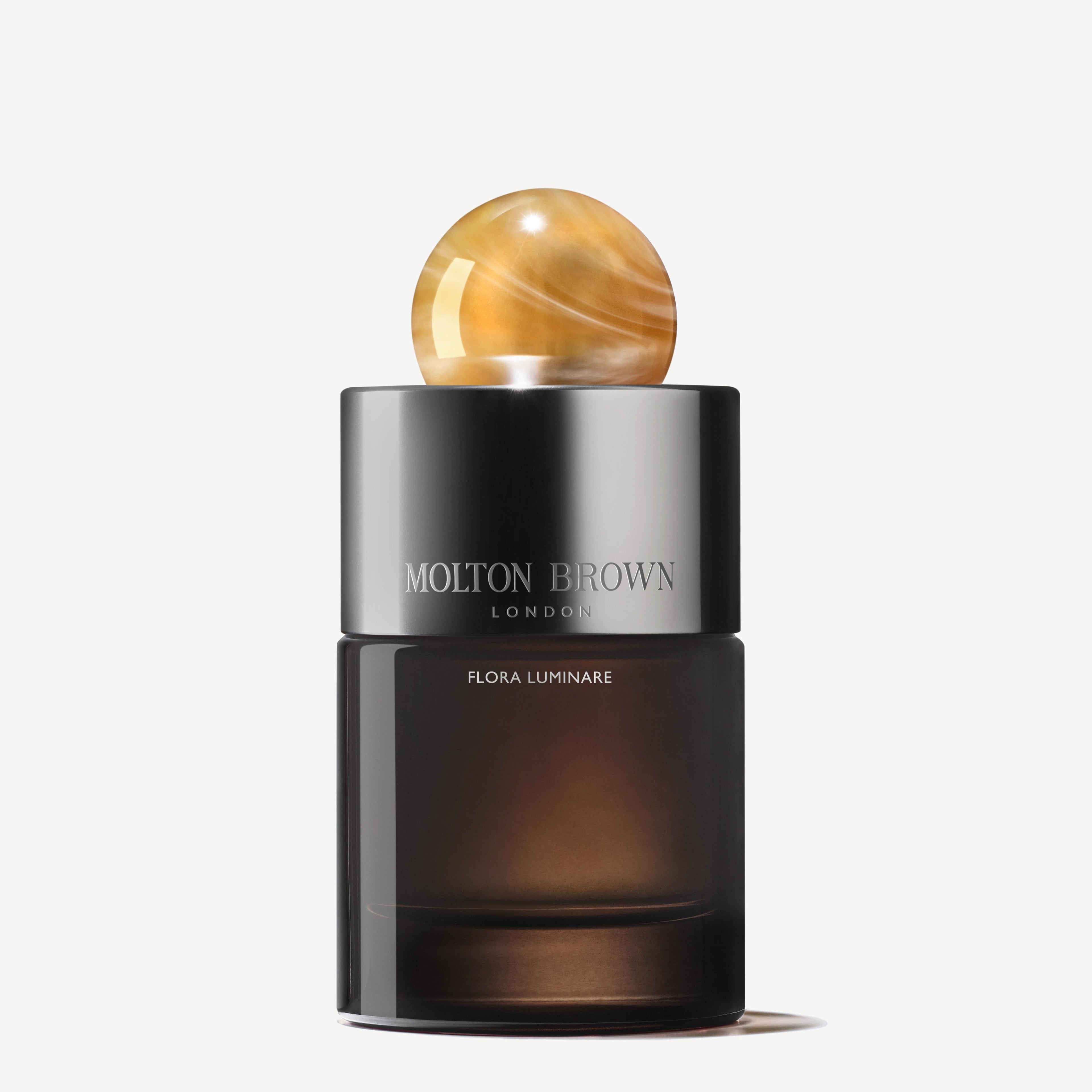 A sleek Flora Luminare Eau de Parfum bottle from MoltonBrown Cyprus, crafted from a refillable glass flacon. The bottle is designed with a dark gradient and is topped with a round, amber-colored cap, set against a plain light background. Its fragrance evokes hints of the warmth of sandalwood.