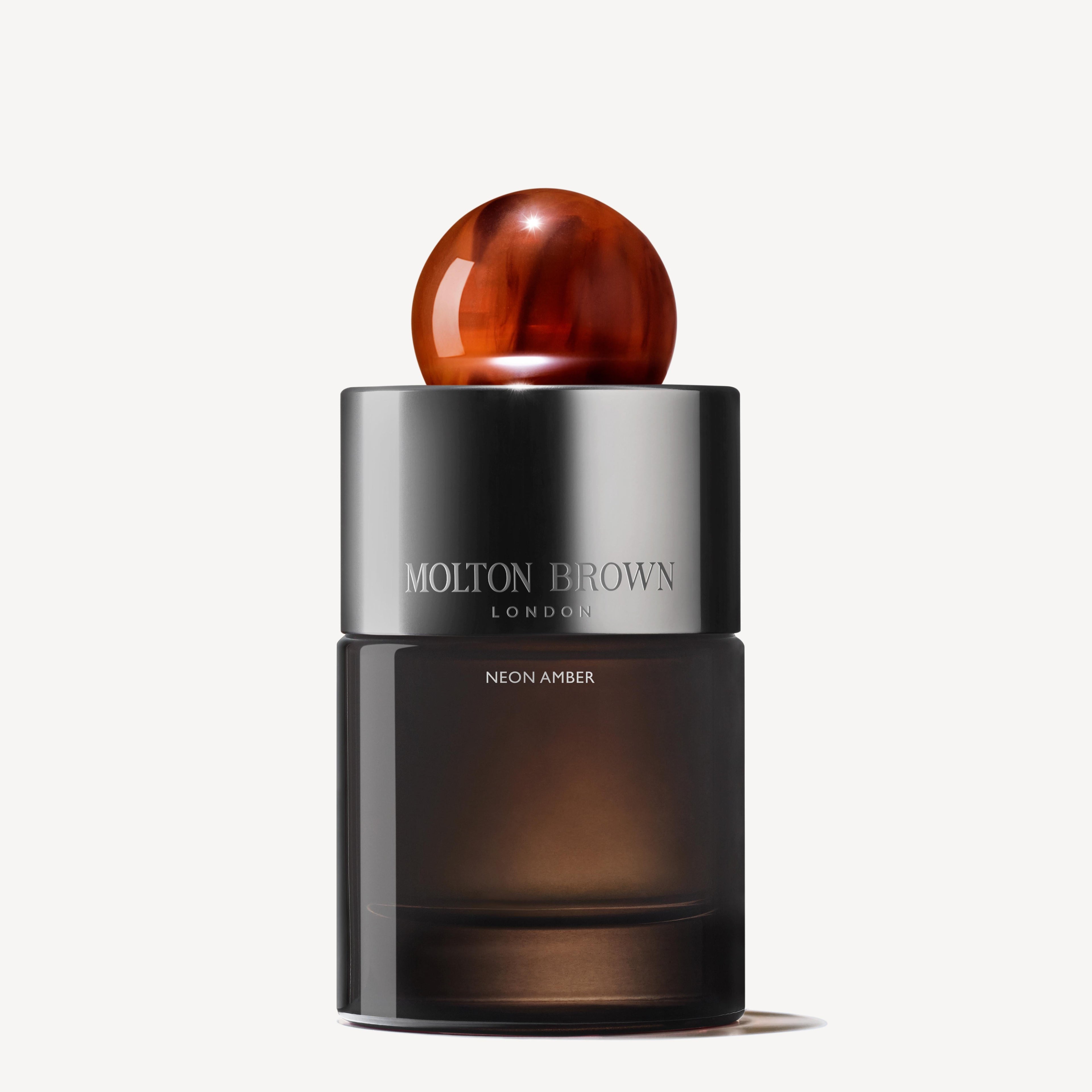 A 100ml bottle of Neon Amber Eau de Parfum by MoltonBrown Cyprus, designed by Jérôme Di Marino, boasts a cylindrical shape that includes a dark glass base and a reflective metallic cap adorned with an amber-like decorative piece. The scent captures the luxurious essence of tonka absolute notes.