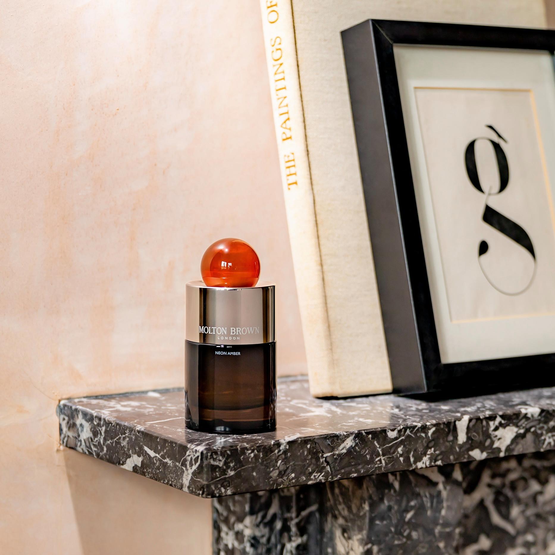 A MoltonBrown Cyprus Neon Amber Eau de Parfum 100ml bottle with a round, red cap sits elegantly on a marble shelf. Beside it lies an artwork featuring a stylized letter g and a hardcover book with the spine partially visible, capturing Jérôme Di Marino's essence and tonka absolute sophistication.