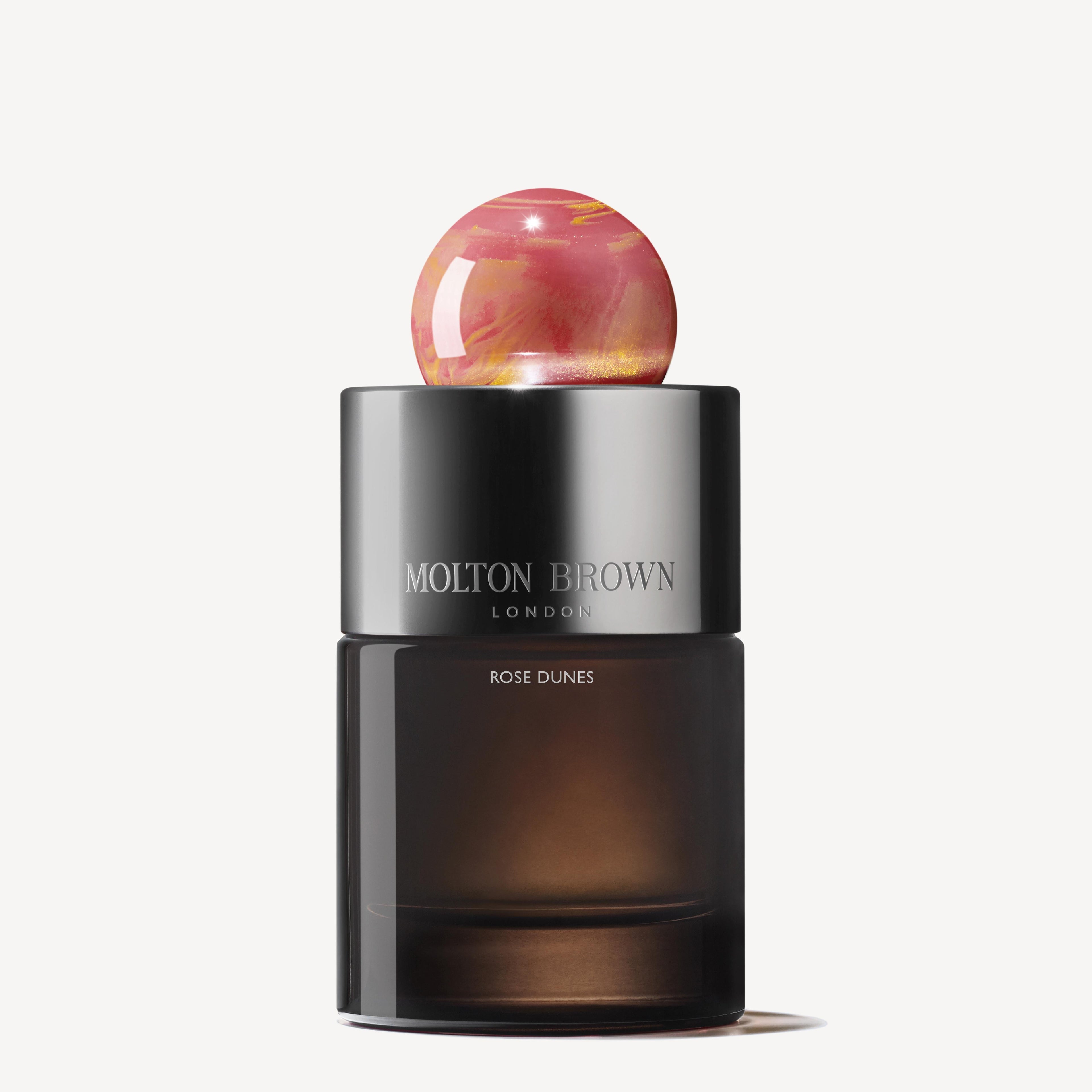 A sleek, dark glass bottle of MoltonBrown Cyprus's Rose Dunes Eau de Parfum 100ml is displayed, featuring a spherical, marbled pink and orange cap. The brand name and product details are printed on the bottle, hinting at the opulent oudh accord within.